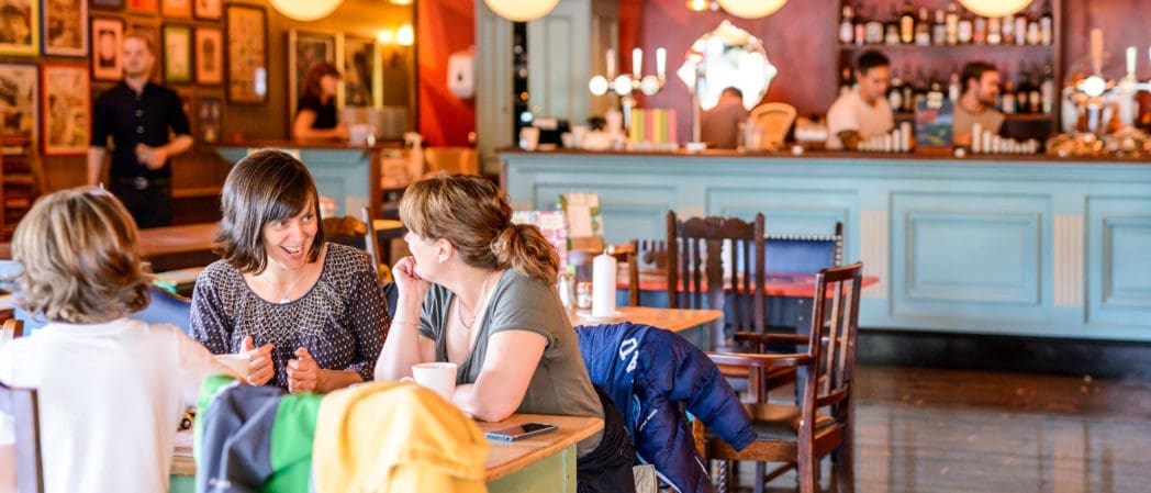 Two women sit at a café table, smiling and chatting over drinks. A child sits nearby as Bolero plays softly in the background, while people work behind a teal counter. Warm lighting and framed artwork create a cozy, lively atmosphere.