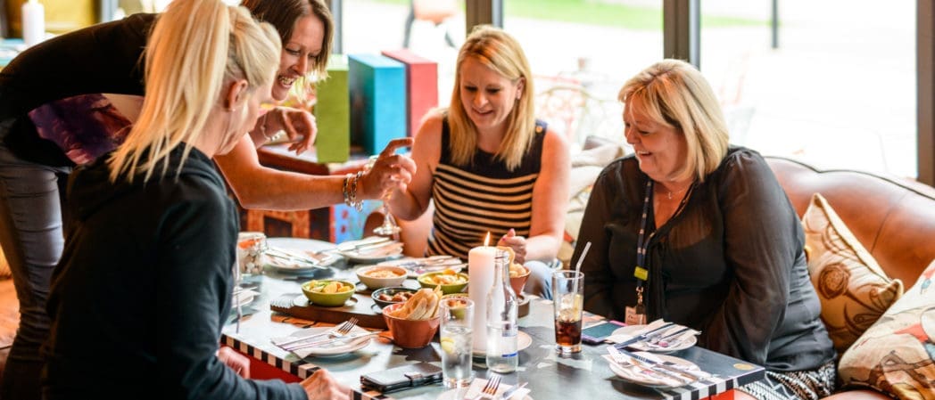 Four women sit around a table in a brightly lit restaurant, laughing and sharing food. A woman in a stylish bolero adds flair to the cheerful scene, as natural light streams through large windows onto plates and drinks.