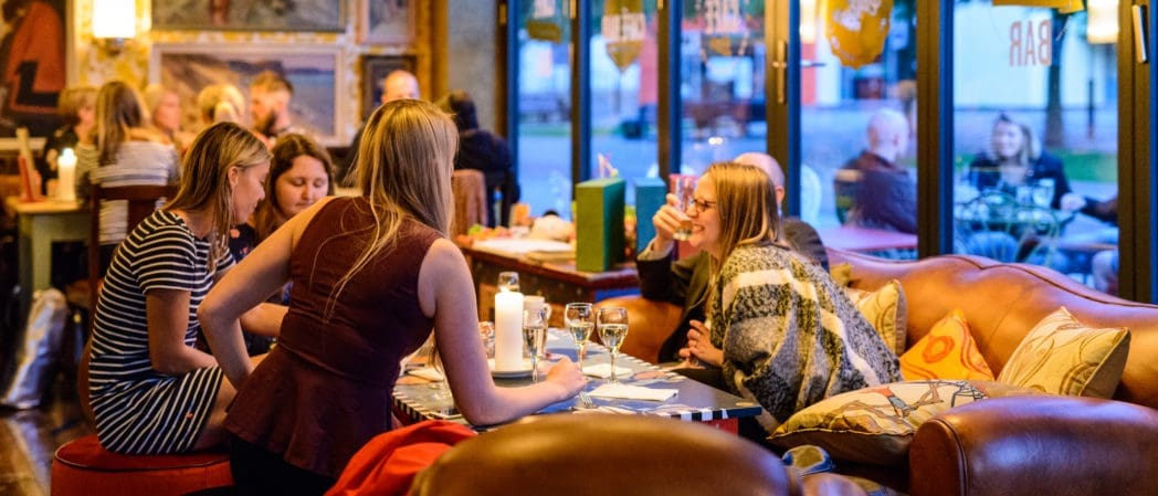 A group of people sit around a table in a cozy, warmly lit restaurant, talking and enjoying drinks as the gentle sounds of Bolero drift through the air, with other diners visible in the background and outside through large windows.