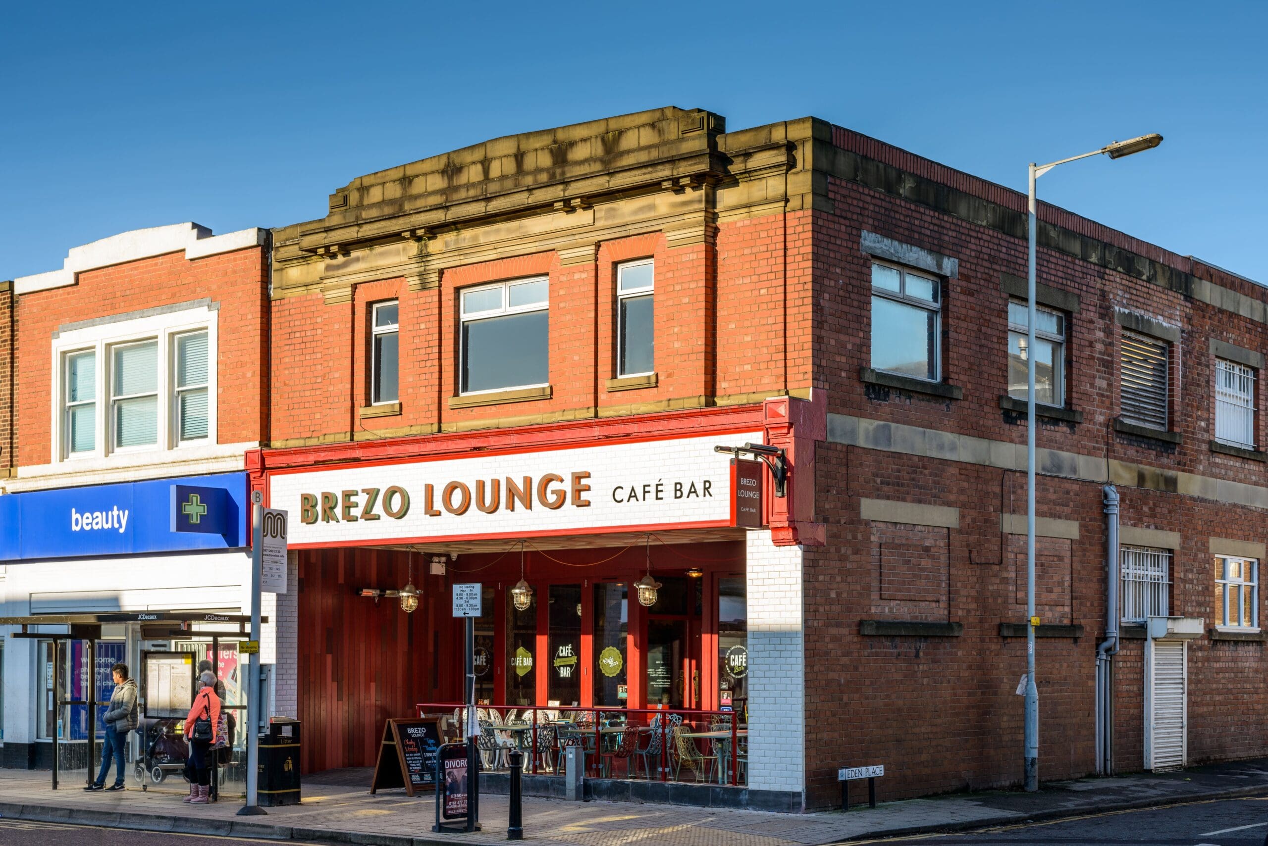 A red-brick corner building houses Brezo Lounge café bar, with outdoor seating and large windows. Next door is a shop labeled "beauty." Several people stroll past Brezo along the sidewalk under a clear blue sky.