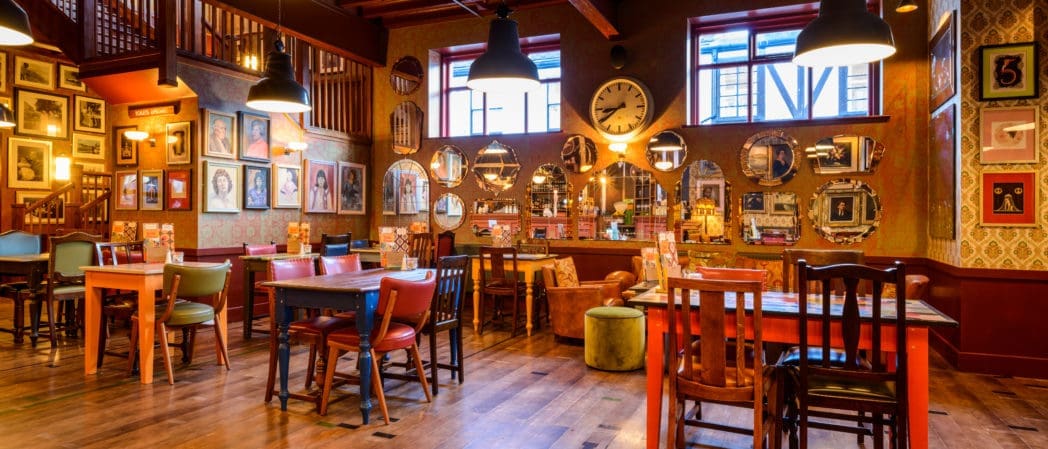 A cozy, eclectic restaurant interior with mismatched wooden chairs and tables, vintage wall art, and warm lighting. Large windows and a prominent clock give Brezo a vibrant character and inviting charm.