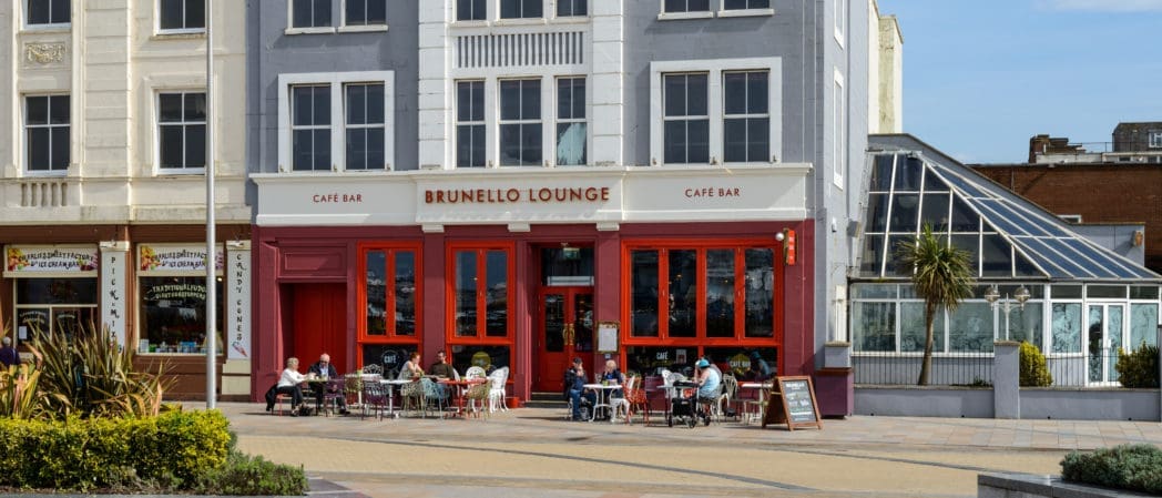 People sit at outdoor tables in front of Brunello Lounge café bar, a gray and red building with large windows. Nearby, other shops and a glass conservatory create a lively scene on this sunny day near Brunello.