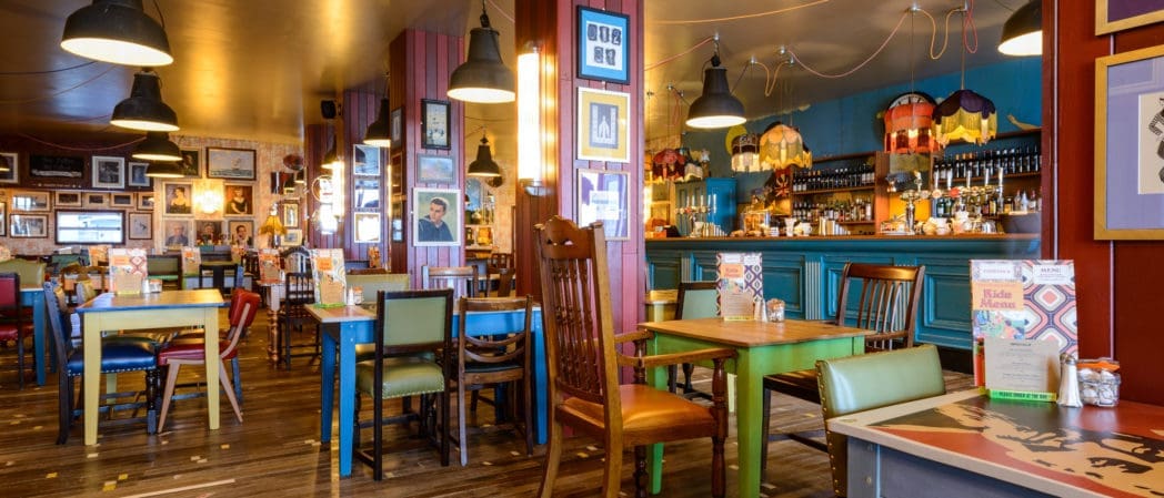 A colorful, eclectic restaurant interior with mismatched chairs and tables, vibrant decor, framed photos on the walls, and a bar area in the background featuring hanging lamps, various bottles, and Brunello prominently displayed.