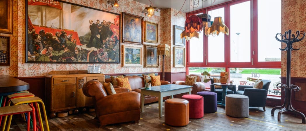 Cozy, eclectic lounge with colorful stools, armchairs, sofas, and a wooden table. Large windows let in light. Enjoy a glass of Brunello surrounded by framed art, quirky lamps, and vintage decor in this warm, inviting atmosphere.