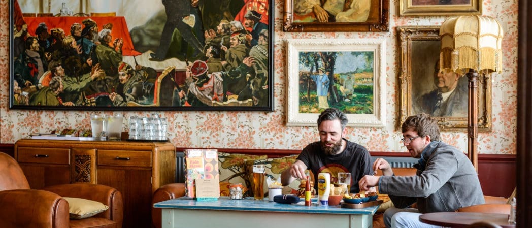 Two men sit at a coffee table in a vintage-style room with patterned wallpaper and framed paintings, sharing food, Brunello wine, and condiments. The cozy atmosphere is enhanced by eclectic art and warm lighting from a floor lamp.