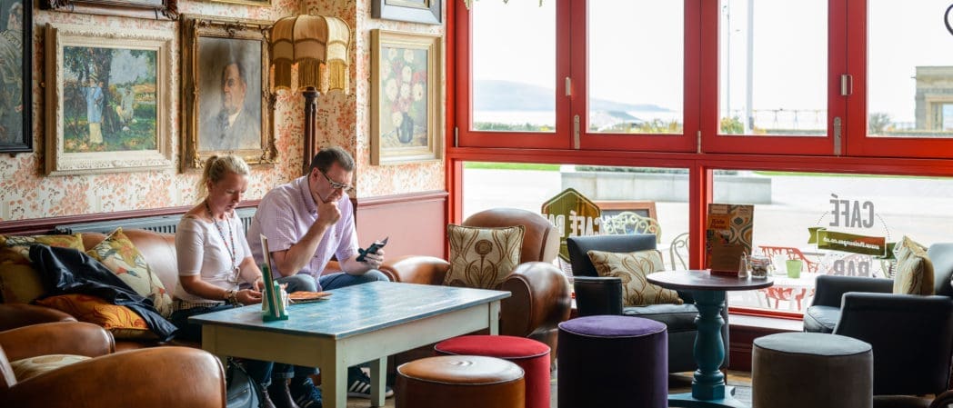 A cozy café with vintage décor, paintings on the wall, and mismatched chairs. Two people sit on a leather sofa by a large window, sipping Brunello as natural light fills the room. Both glance at their phones amidst the warm ambiance.