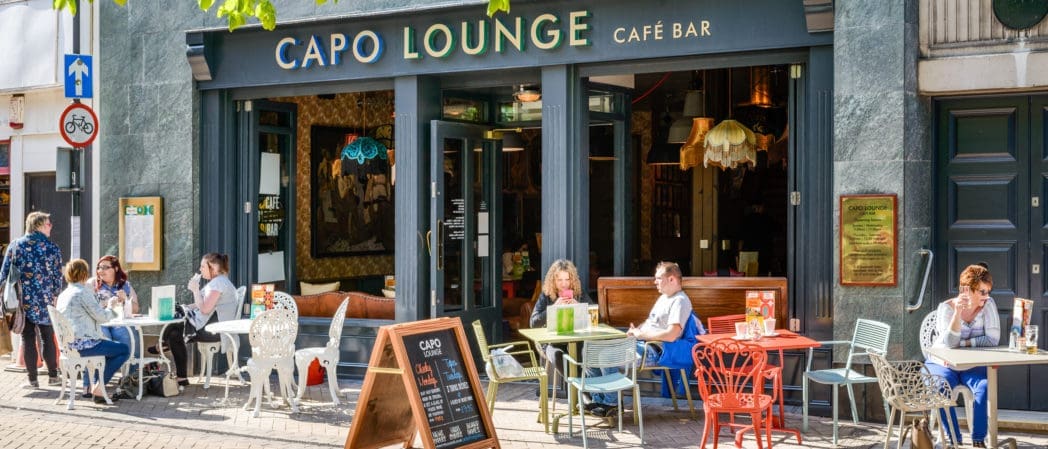 People relax at colorful tables outside Capo Lounge café bar on a sunny day, enjoying drinks and conversation. Capo features large windows, decorative signs, and eclectic chairs lining the lively sidewalk.