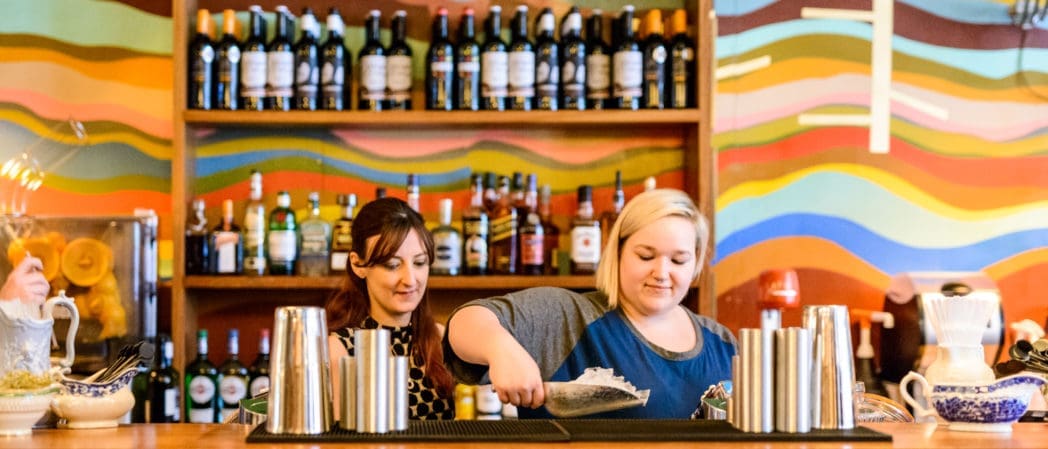 Two women work behind the colorful Capo bar, one pouring ice into a glass and the other preparing drinks. Bottles of alcohol are neatly arranged on shelves, with a bright, wavy mural on the wall.