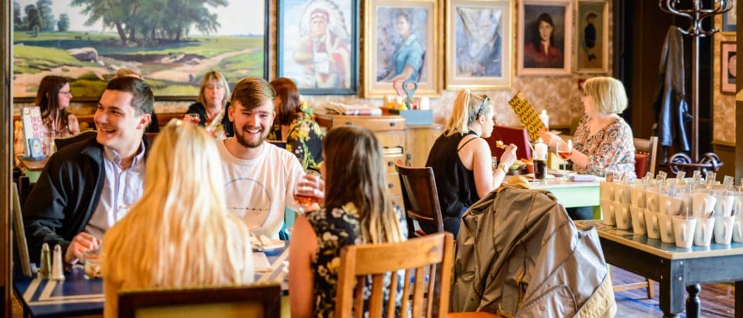 A lively café scene with groups of people chatting and dining at tables. Colorful paintings, portraits, and a vintage Capo coffee machine create a cozy, artistic atmosphere. Cups and tableware are visible in the foreground.