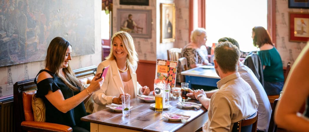 A group of people sit around a wooden table in Capo, a cozy, art-filled restaurant, smiling and chatting. One woman is taking a photo on her phone while others enjoy drinks and conversation. Other diners are visible in the background.