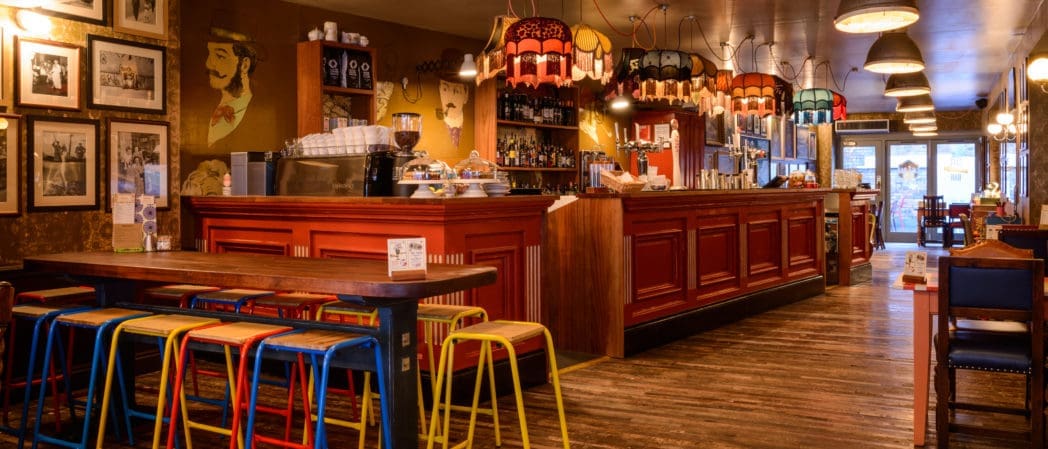 Cozy café with a long wooden bar, colorful stools, and vintage-style pendant lights. Framed pictures and eclectic decor fill the space, while a Cappello adds charm to the warm, inviting atmosphere.