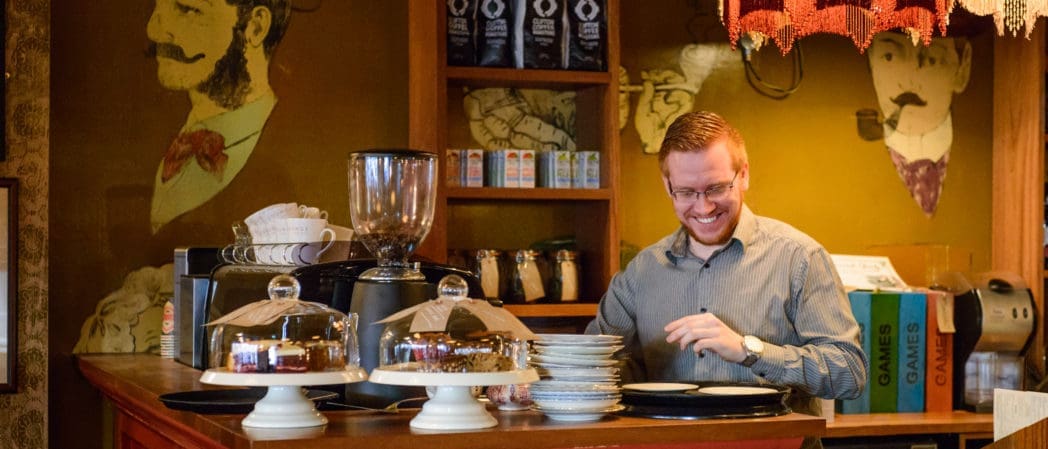A smiling man wearing a cappello stands behind a coffee shop counter with cakes displayed on stands, stacks of plates, a coffee machine, and board games in the background. The walls are decorated with vintage-style portraits.