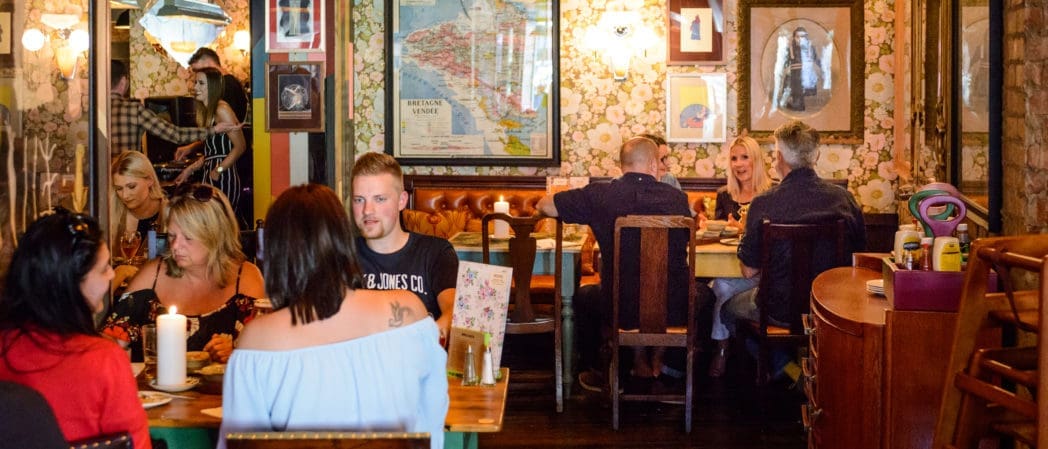 Several groups of people sit at wooden tables in Carnero, a cozy, warmly lit restaurant with floral wallpaper, framed art, and vintage decor, chatting and enjoying their meals.