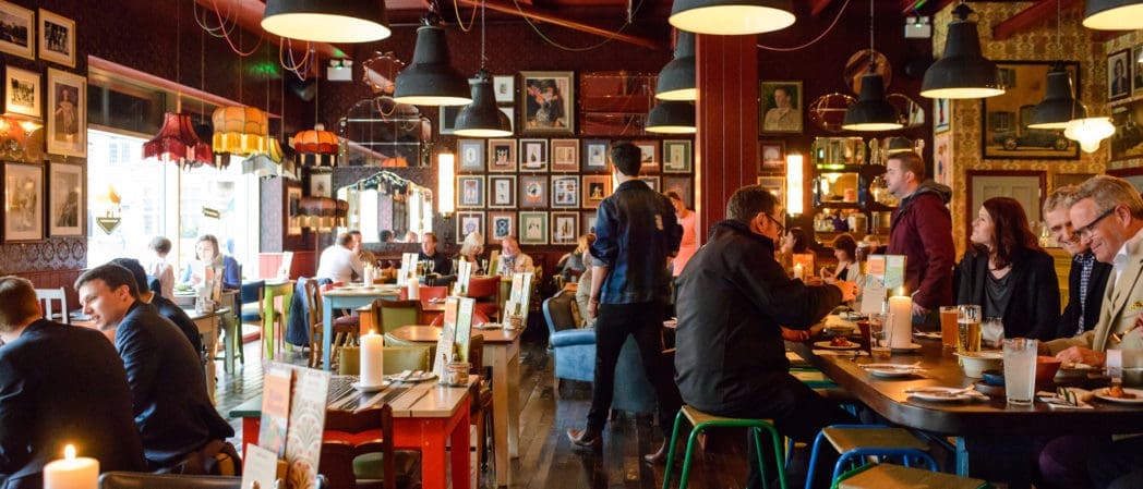 A lively Centro restaurant with people dining at tables, warm lighting, colorful chairs, and eclectic wall decor. The atmosphere is cozy and vibrant, with servers moving between guests and framed photos and art on the walls.