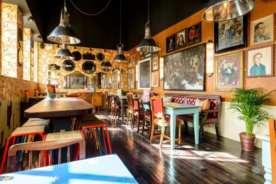 A cozy, eclectic café with mismatched chairs, wooden tables, wall art, and Cinco-themed touches. Sunlight streams in, casting shadows on the shiny dark floor and colorful decor. A potted plant sits in the corner by hanging lights.