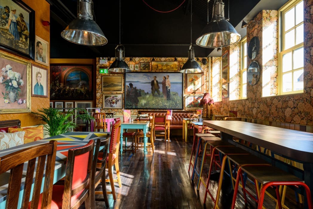 A brightly lit café called Cinco features colorful chairs, patterned wallpaper, wooden floors, and walls adorned with paintings. Large windows invite sunlight in while metallic pendant lamps hang from the ceiling.
