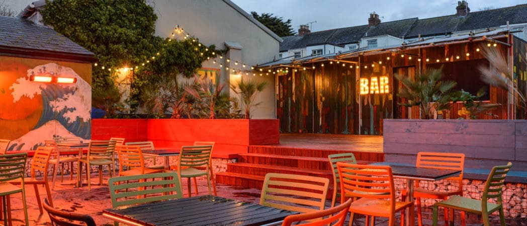 Outdoor bar area with colorful chairs and tables, warm string lights overhead, a mural of a wave, palm plants, and a wooden Concho-style bar with a glowing "BAR" sign, set against houses and the evening sky.