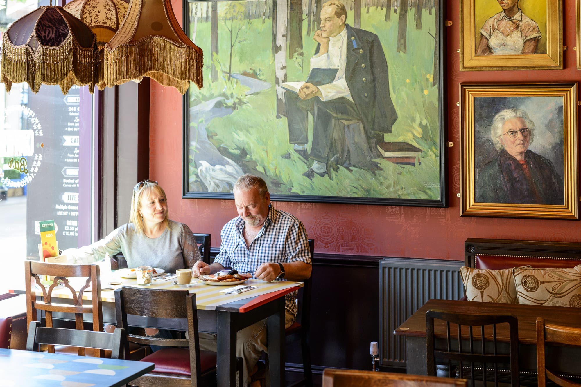 A man and woman sit at a table in a cozy Dorado café, enjoying food and drinks. Large portraits and artwork decorate the warmly lit red walls as sunlight streams through the window onto their table.