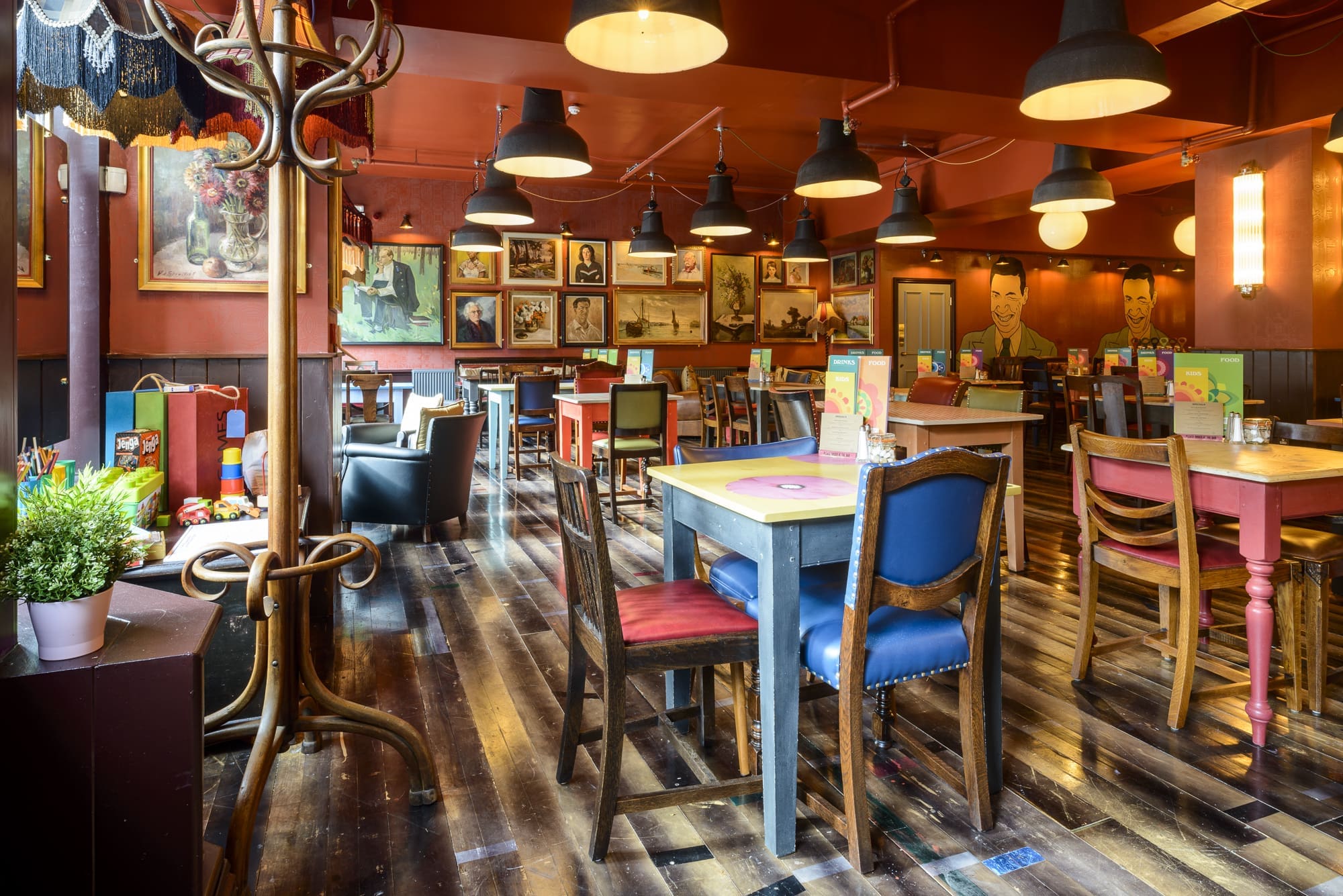 A cozy, colorful café interior with eclectic furniture, wooden floors, framed art on rich Dorado-red walls, and empty tables set for guests under warm, hanging lights.