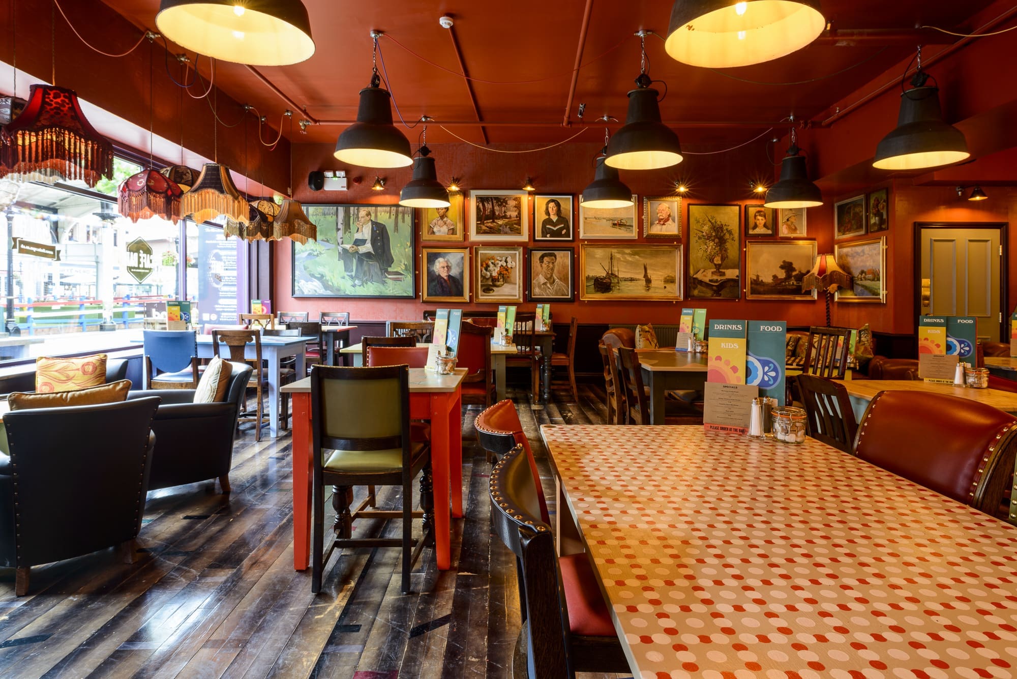 A cozy, eclectic cafe with Dorado-patterned tables, mismatched chairs, warm lighting, and framed artwork covering the walls. Large windows let in natural light, creating a welcoming atmosphere.