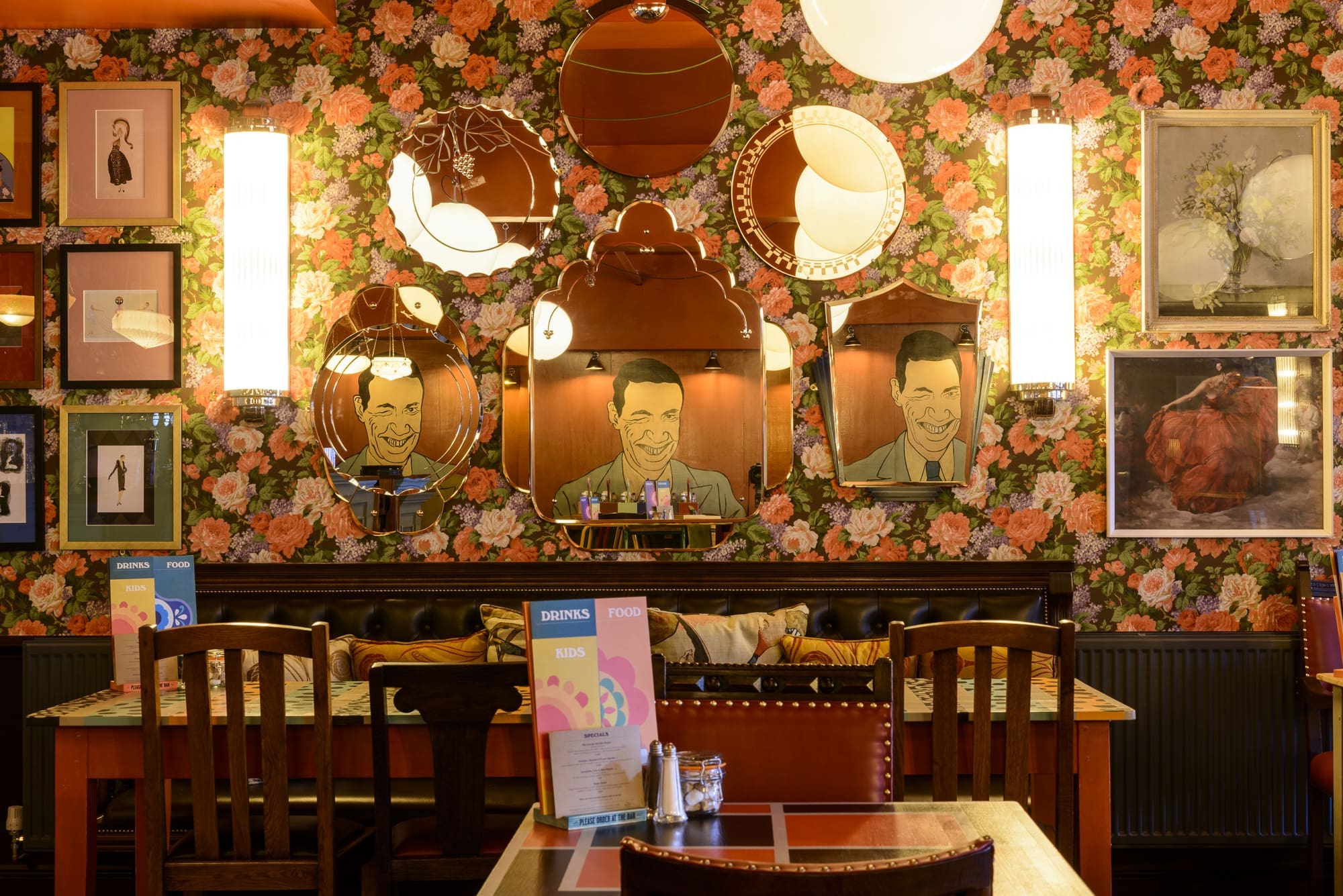 A cozy Dorado restaurant booth with floral wallpaper, decorative mirrors featuring cartoon faces, framed art on the walls, and menus on the table, all warmly lit by wall sconces.