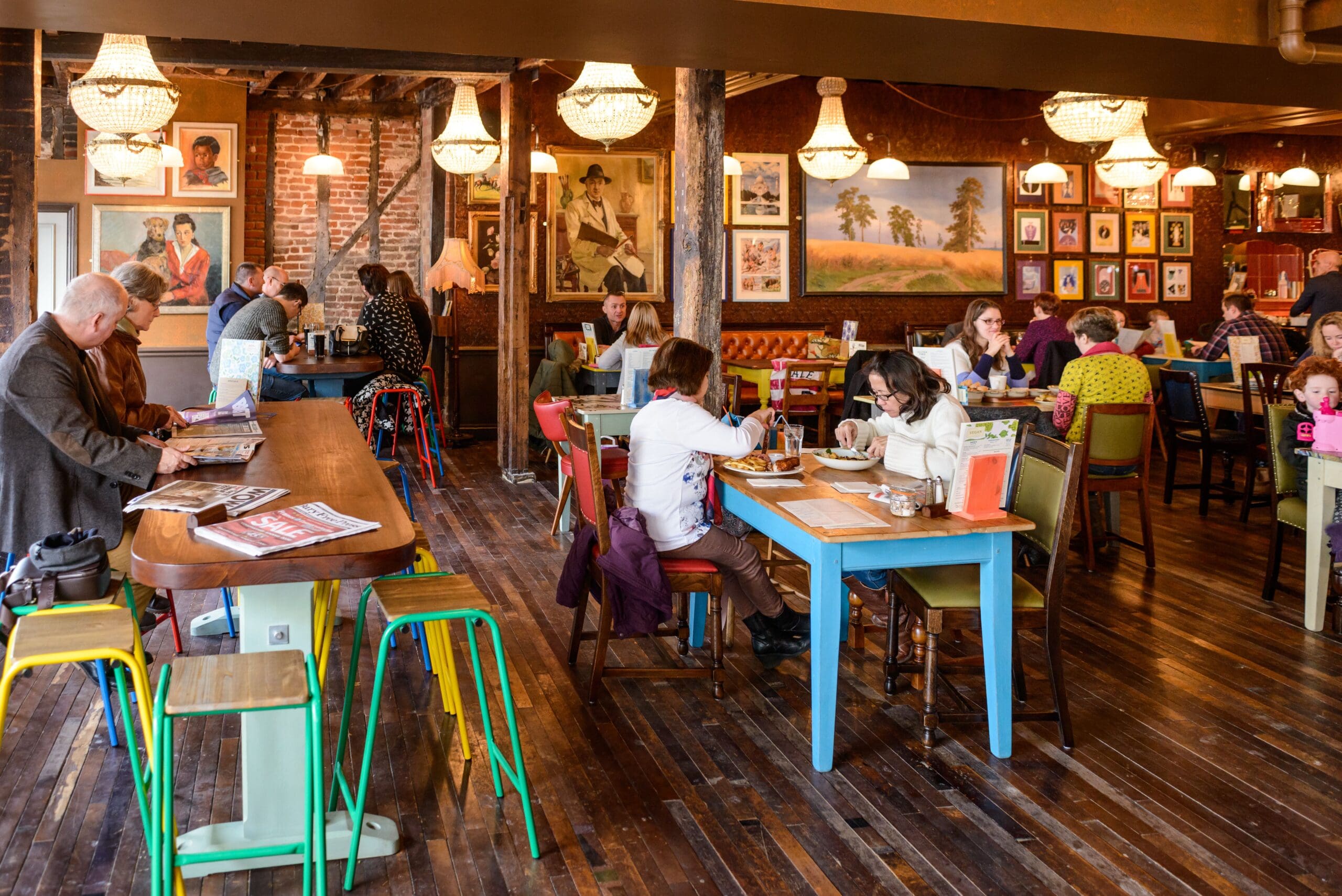 People dine and chat in Edmundo, a cozy, colorful café with wooden floors, art-covered brick walls, and hanging chandeliers. Some enjoy meals at tables, while others read or work at the communal high table.