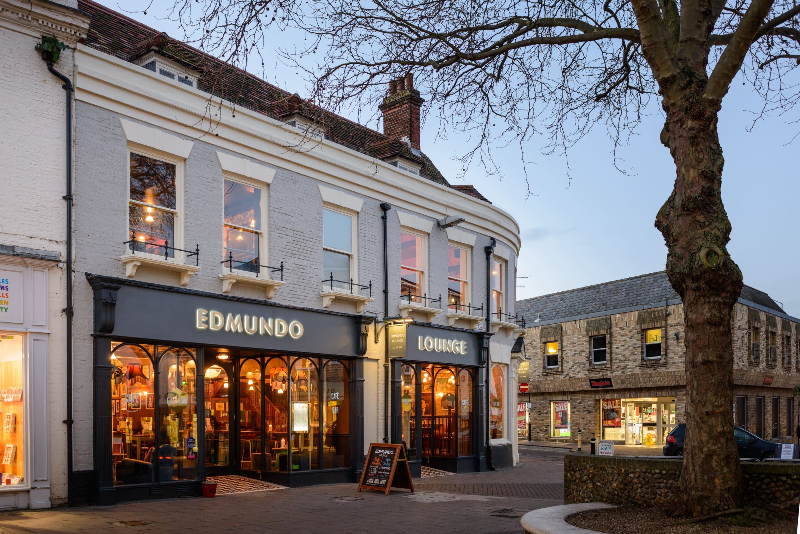 A street view of Edmundo Lounge, a cozy cafe-bar with large arched windows and warm lighting; Edmundo’s outdoor signage stands out amid nearby buildings, a tree, and shops visible at dusk.