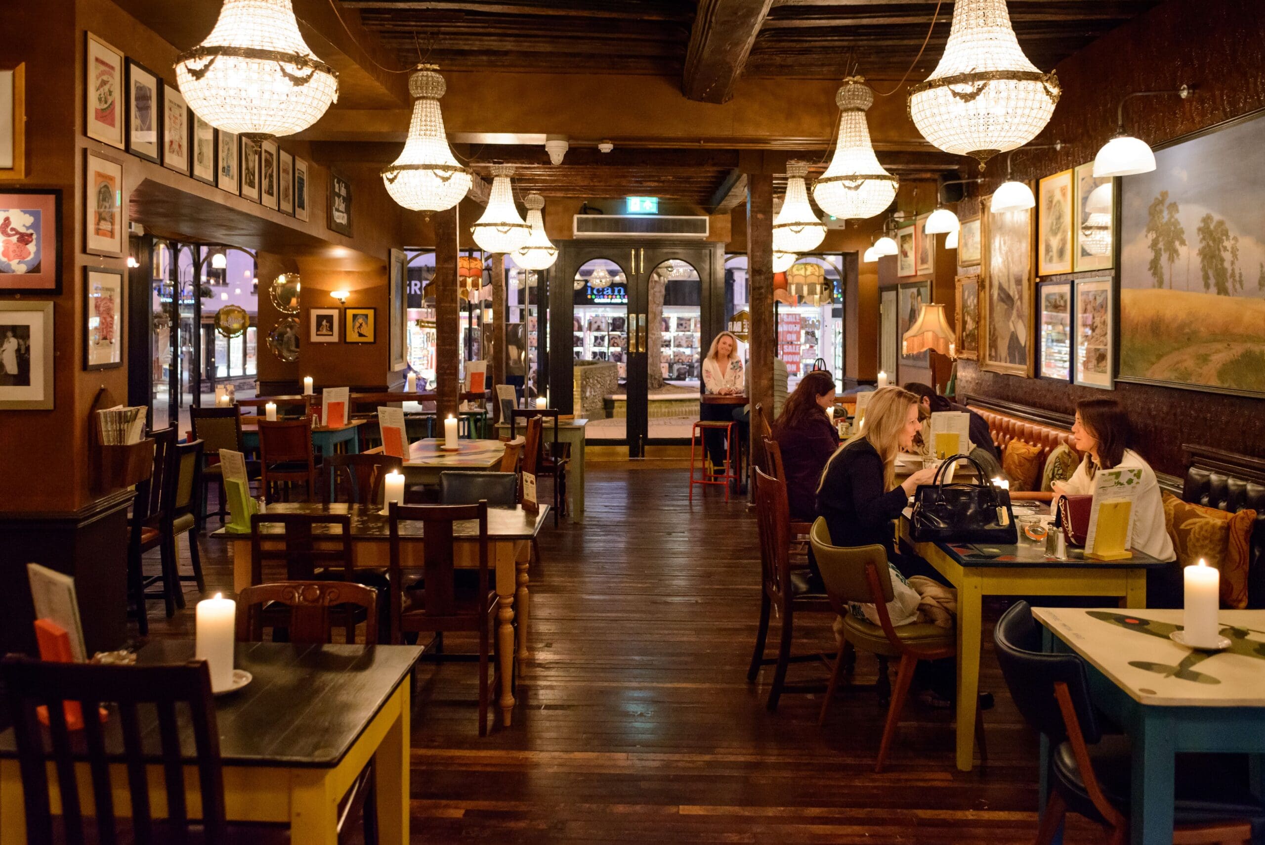 A cozy, warmly lit restaurant with wooden floors, vintage chandeliers, colorful tables, and framed art on the walls sets the scene at Edmundo. Candles add a relaxed, inviting atmosphere as guests dine and chat inside.