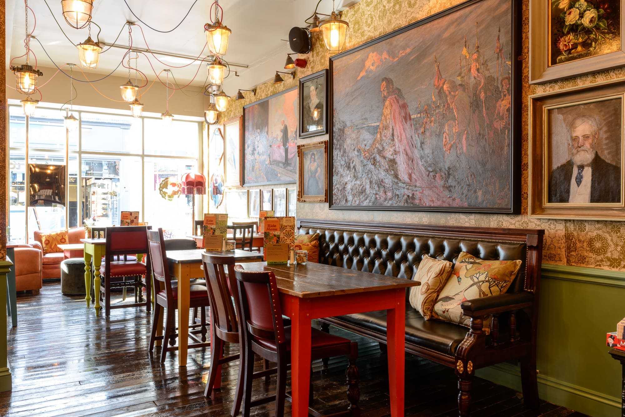 Cozy café in Faro with leather sofa, red tables, and mismatched chairs. Large paintings and portraits hang on patterned wallpaper. Sunlight streams through large windows, illuminating the eclectic, vintage decor.
