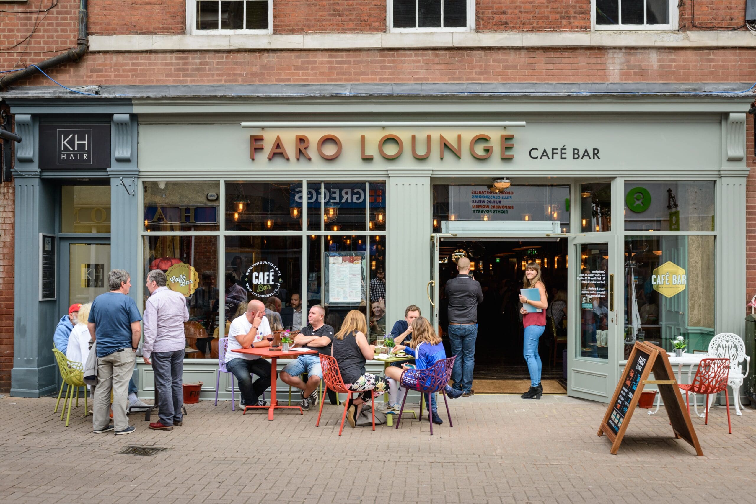 People sit and stand outside Faro Lounge café bar on a bustling pedestrian street, chatting and enjoying drinks. Faro’s large windows, colorful tables and chairs, and welcoming atmosphere make it the perfect lively spot to relax with friends.