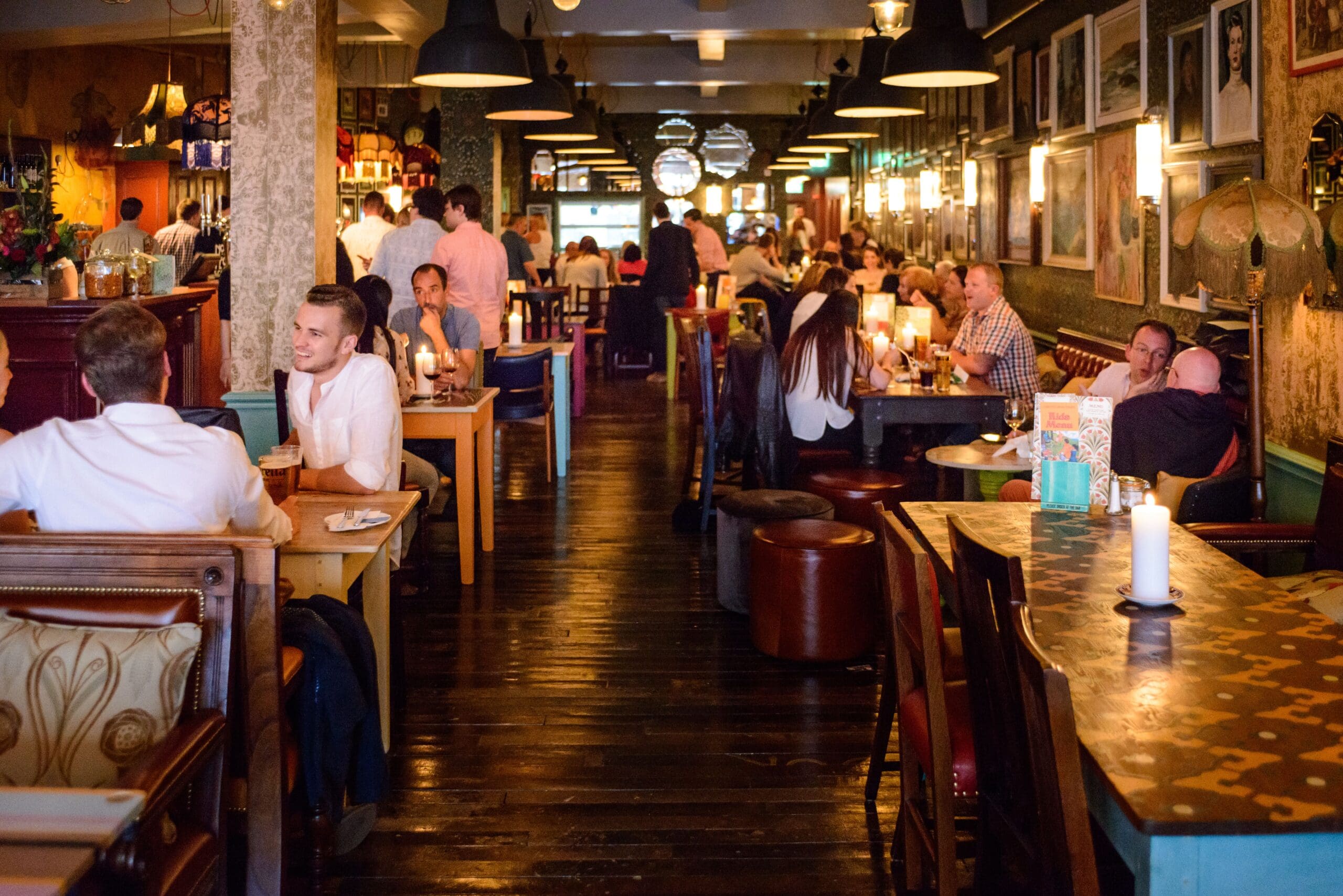 A lively restaurant in Faro with people dining and chatting at tables, warm lighting, wooden floors, and walls adorned with framed pictures and art. Candles flicker on the tables, enhancing the cozy, welcoming atmosphere.