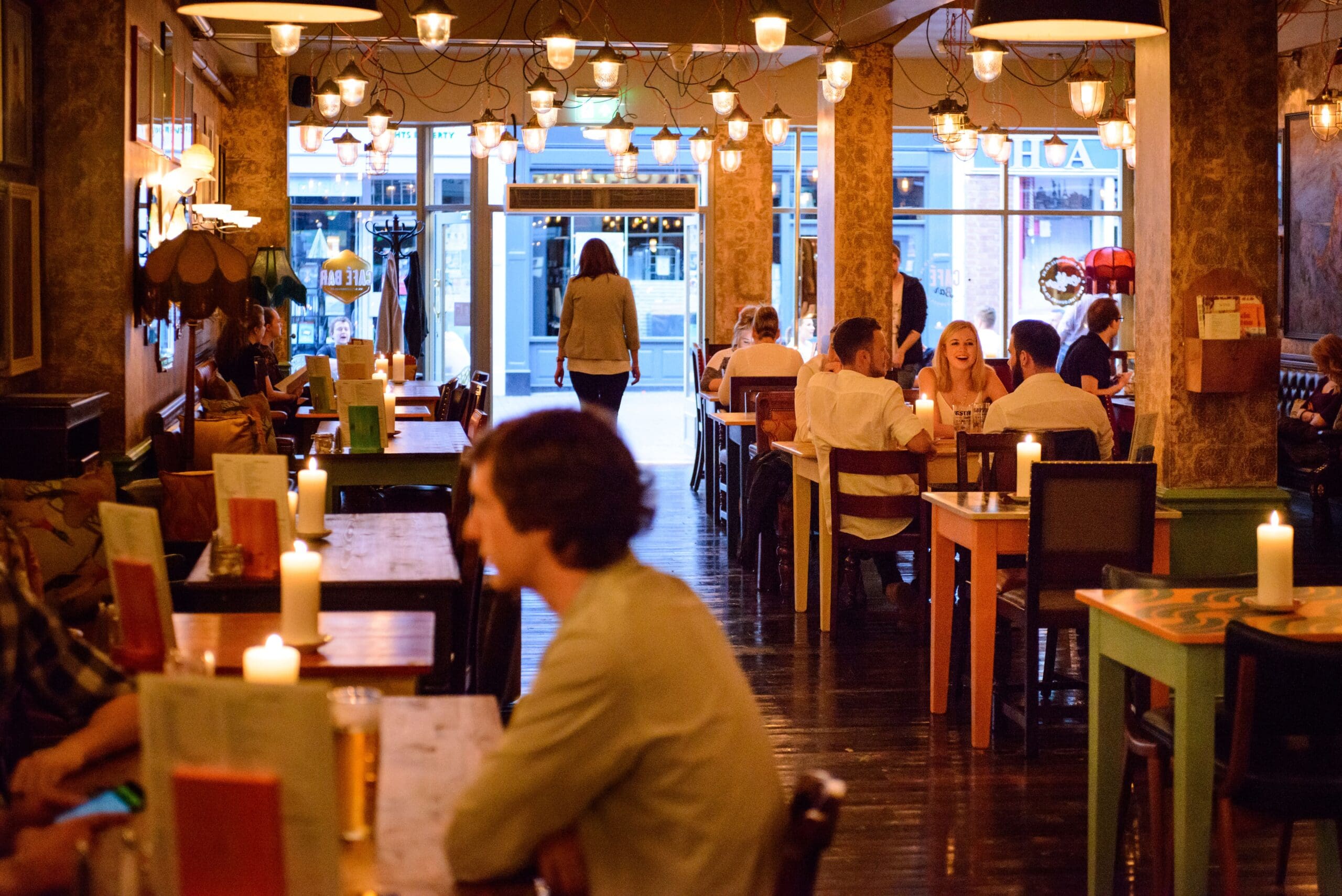 A warmly lit restaurant in Faro filled with people dining at tables. Candles are on the tables, and a person is walking toward the entrance. The overall atmosphere appears cozy and inviting.