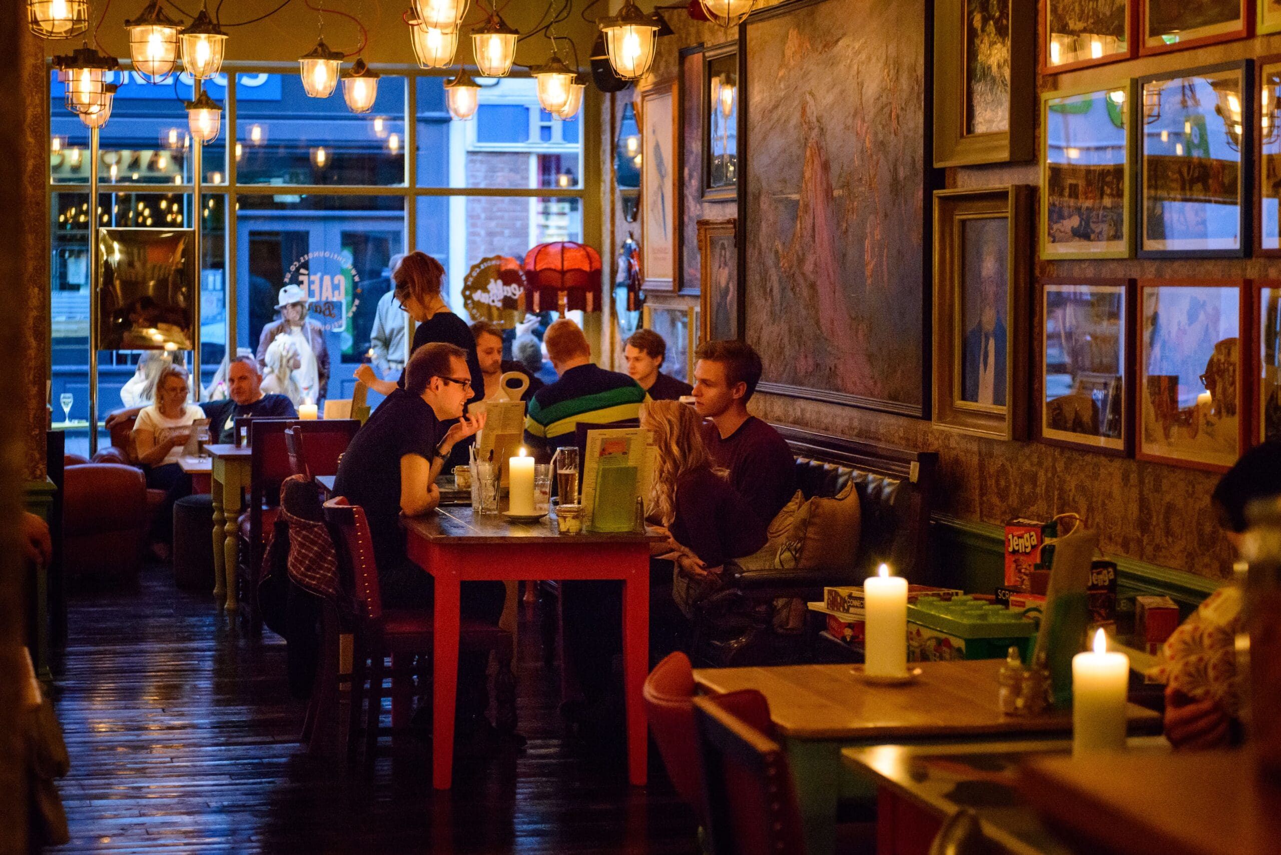 A cozy, dimly lit restaurant in Faro with people dining at tables, warm hanging lights, candles on the tables, eclectic wall art, and large windows revealing more people outside. The atmosphere is lively and intimate.