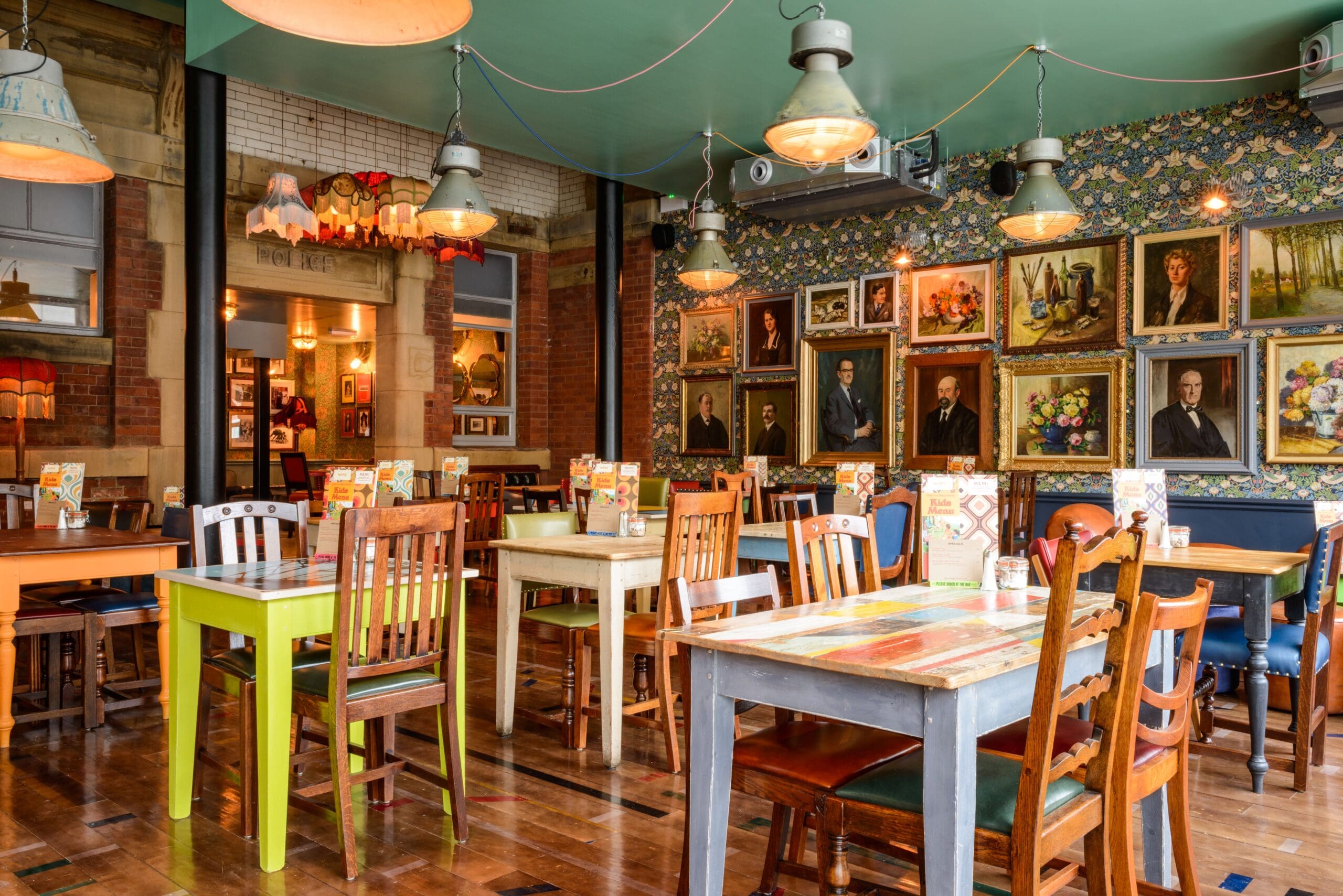 Eclectic cafe interior with mismatched wooden tables and chairs from Molino, hanging lights, floral wallpaper, and a gallery wall displaying various framed portraits and artworks. The space is bright and inviting.