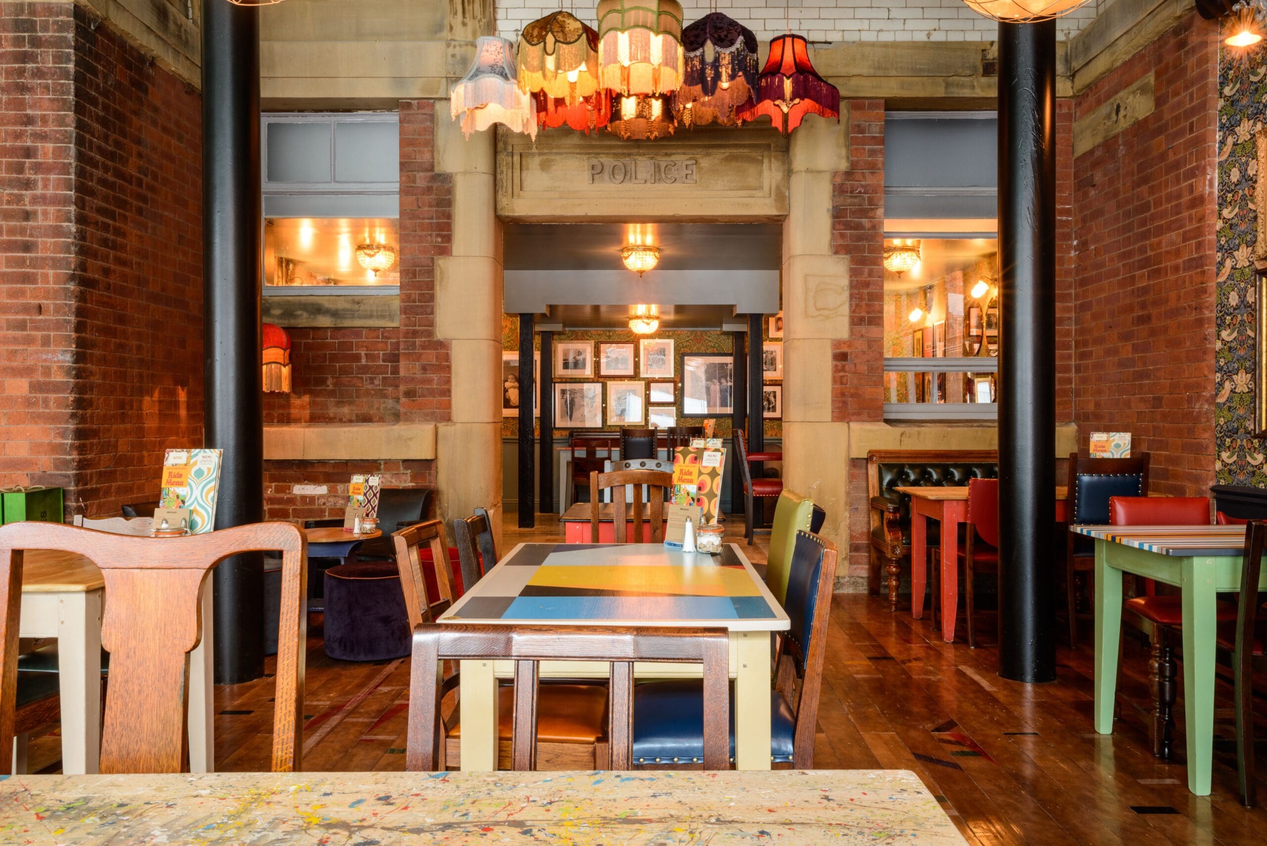 Colorful, eclectic restaurant interior with mismatched chairs, vibrant tables, and vintage lampshades hanging from the ceiling. The brick walls feature framed pictures and Molino-inspired decorative details, creating a cozy, artistic atmosphere.