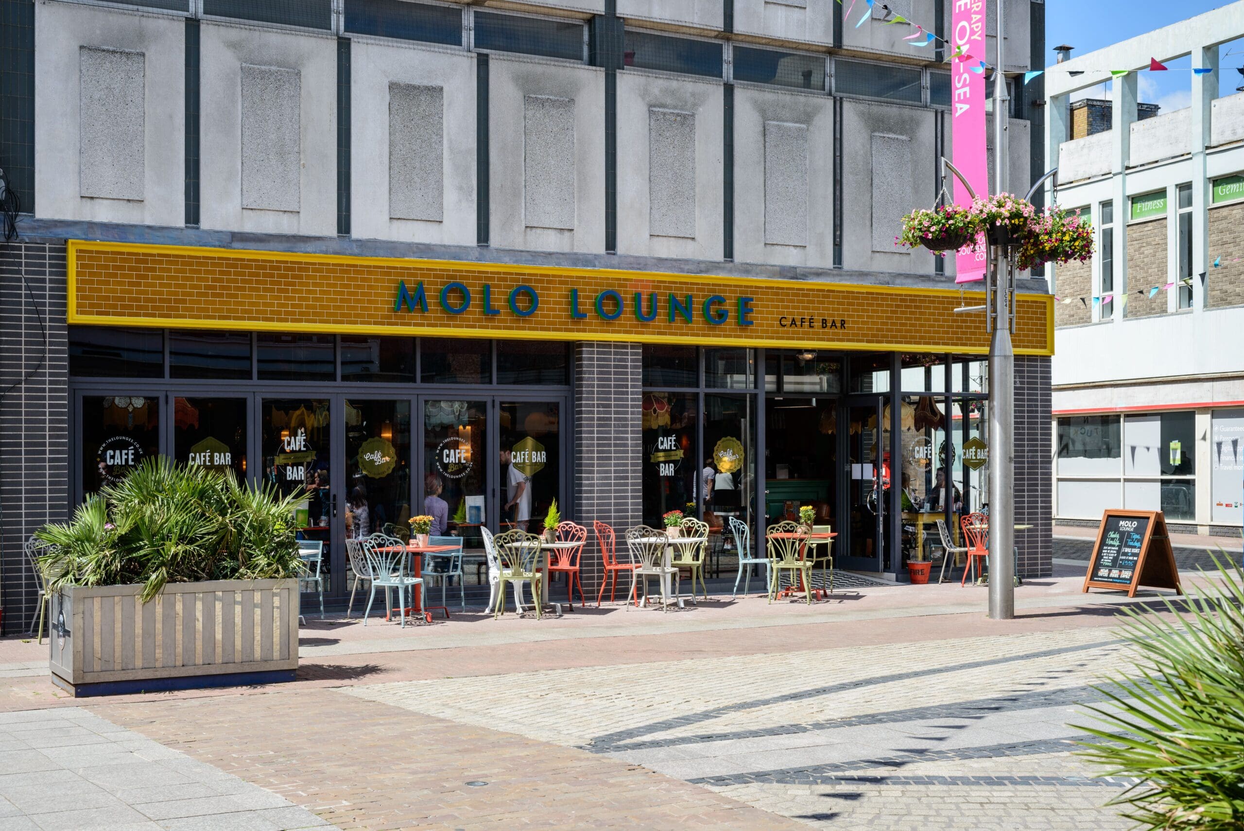 Street view of Molo Lounge café bar with bright yellow Molo signage, outdoor seating featuring colorful chairs and tables, potted plants, and a sandwich board on a paved pedestrian area under a clear sky.