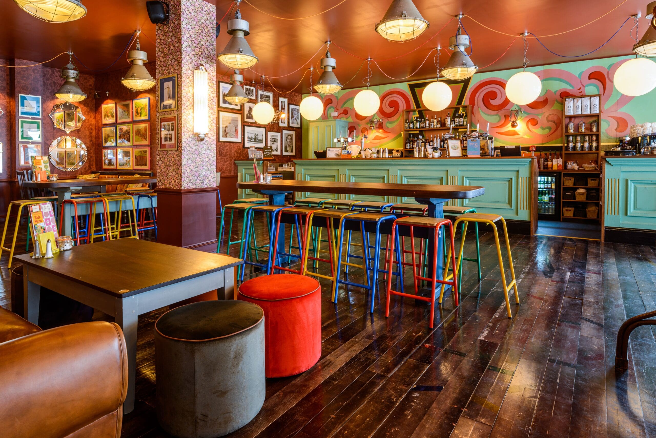 A vibrant bar with colorful Molo stools, round poufs, wooden tables, and eclectic decor. Bright hanging lights, bottles on display, and a mural with swirls create a lively atmosphere.