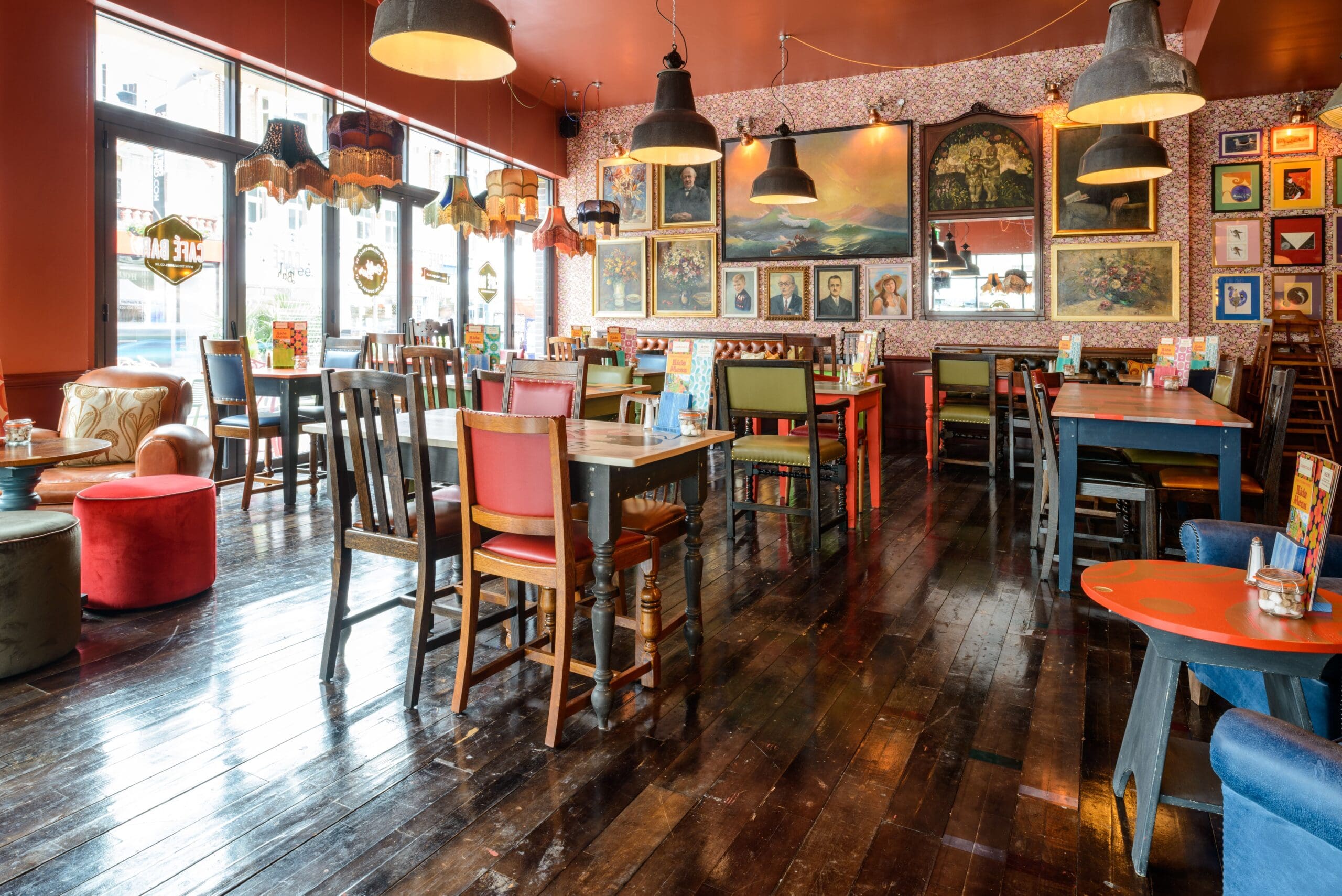 A cozy, eclectic restaurant with Molo-inspired colorful mismatched chairs, wooden tables, hanging lamps, and framed artwork all over the patterned walls. Sunlight streams in through large windows onto the polished wooden floor.
