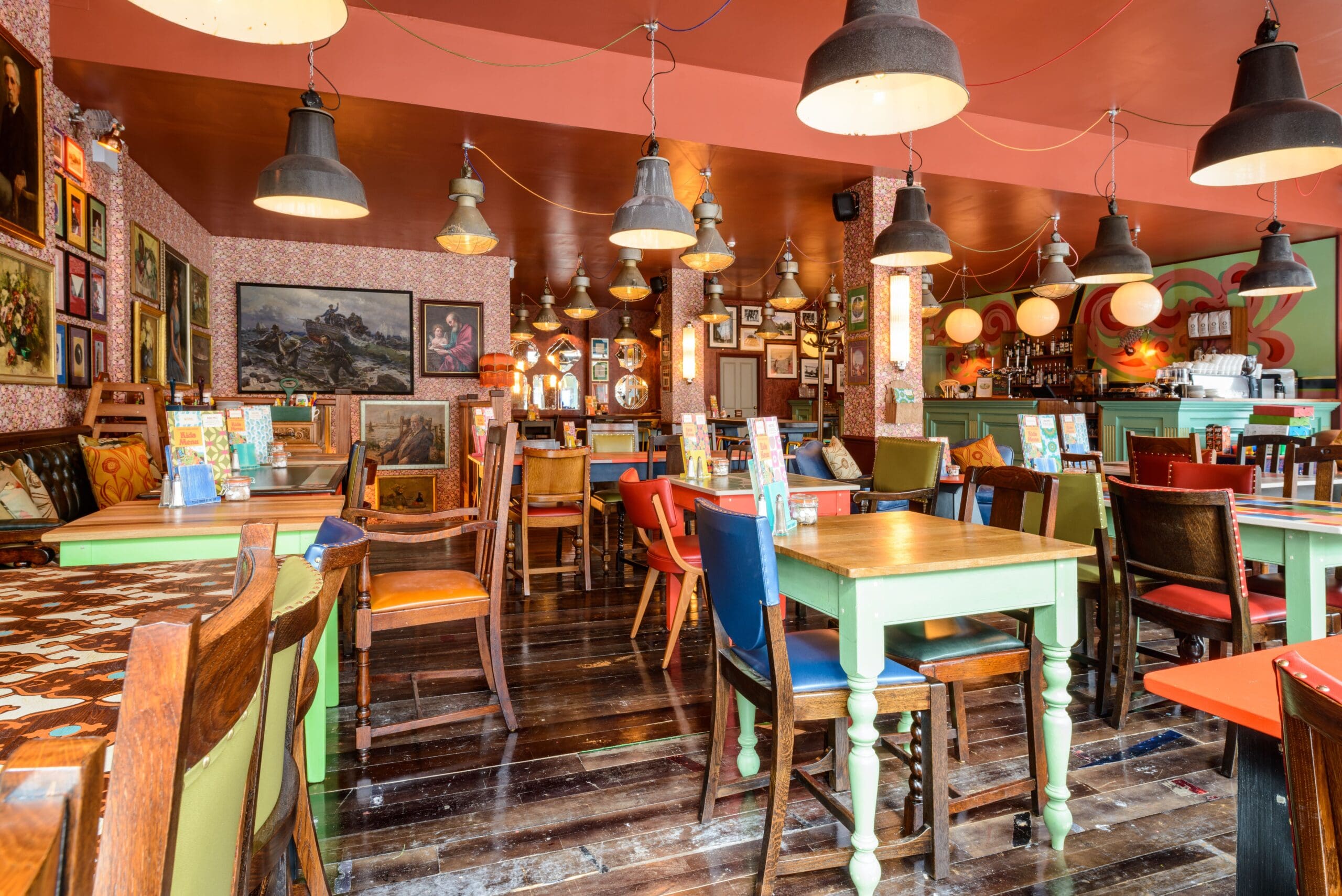 A cozy, eclectic café with colorful wooden tables—some crafted from Molo designs—and mismatched chairs, warm lighting, patterned wallpaper, and various paintings and decorations on the walls. The space appears inviting and vibrant.