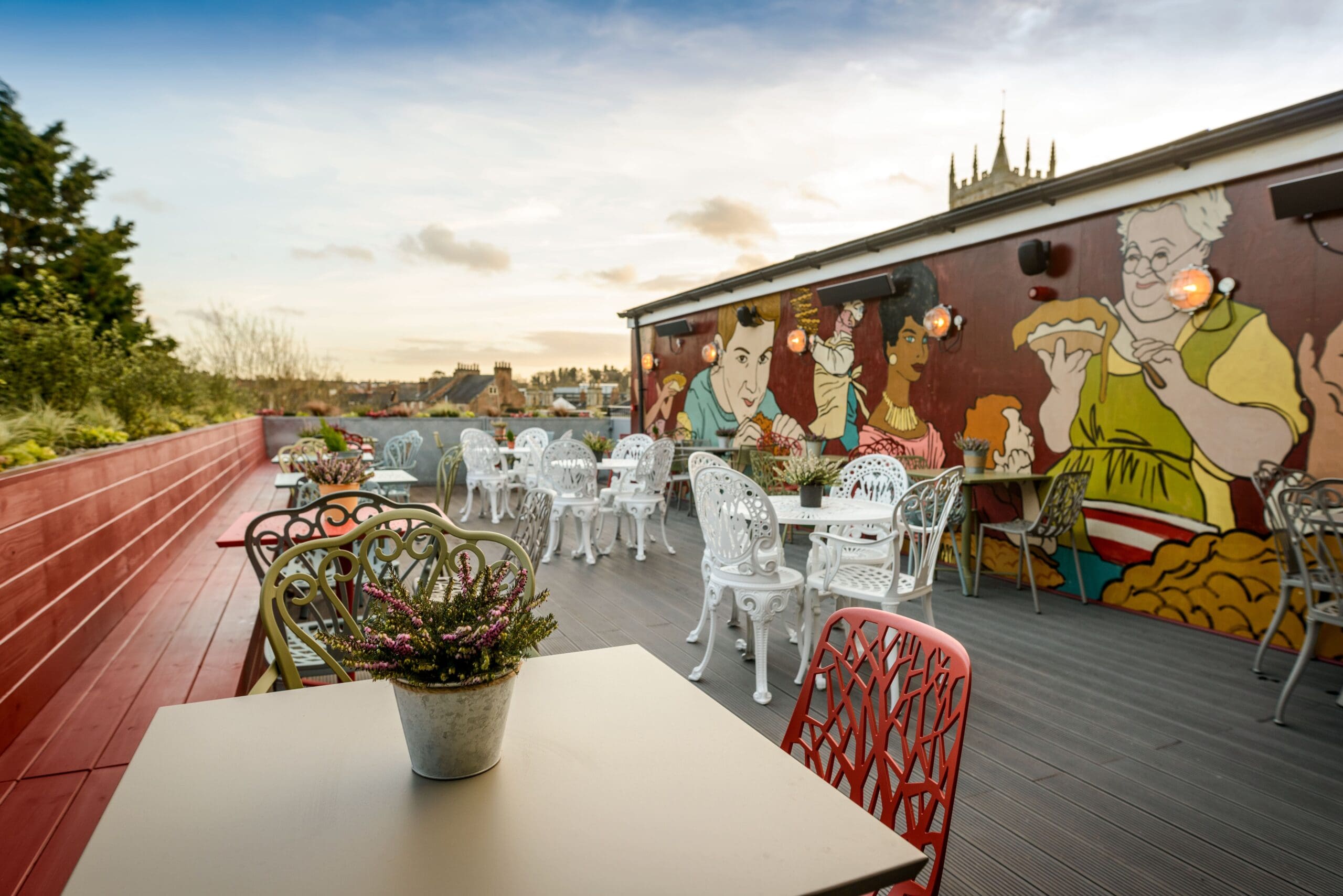 A rooftop patio with decorative metal chairs and tables, potted plants, and a colorful mural of people eating on the wall, with city buildings, the Montero skyline, and a church spire visible in the background.