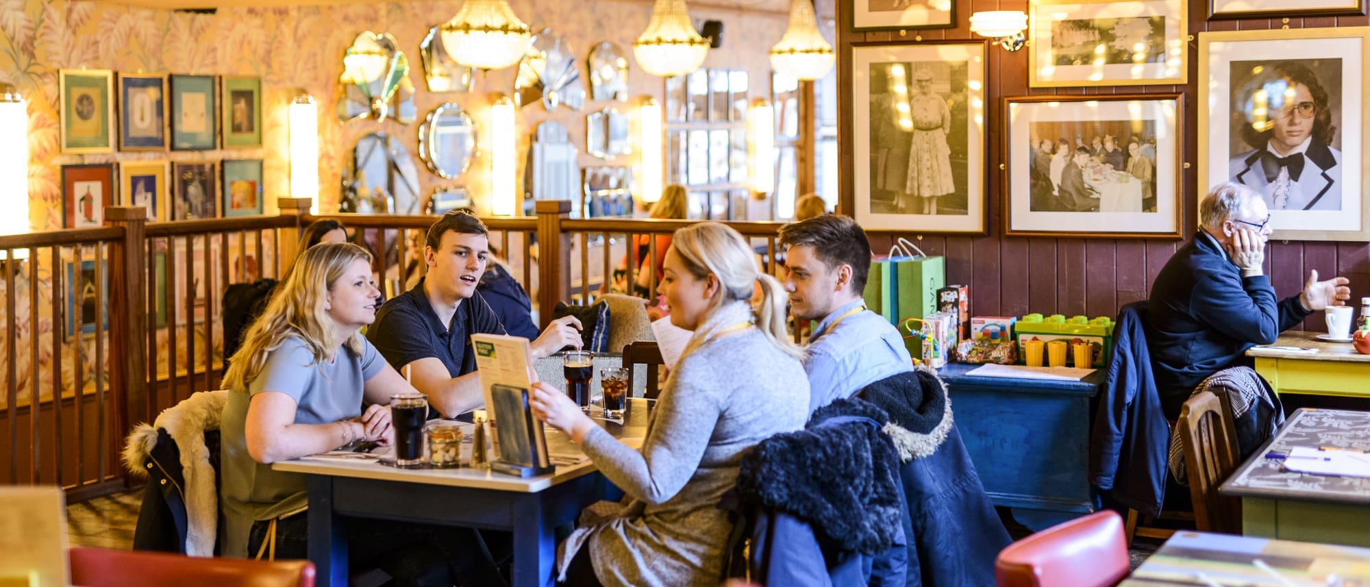 Four young adults sit at a restaurant table in Paramo, chatting and looking at menus, while an older man sits alone nearby. The restaurant features warm lighting, framed photos, and patterned wallpaper on the walls.