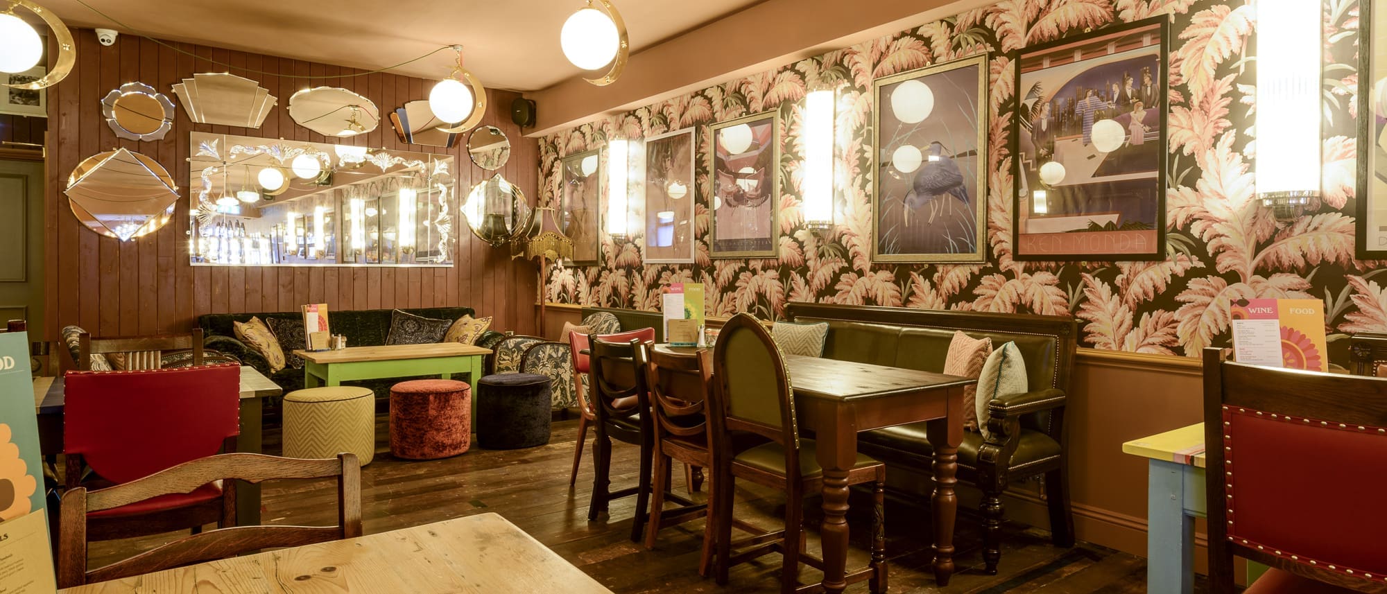 A cozy Paramo café with eclectic vintage decor, patterned wallpaper, round mirrors, cushioned seating, wooden tables, and warm ambient lighting. Menus are placed on the tables.
