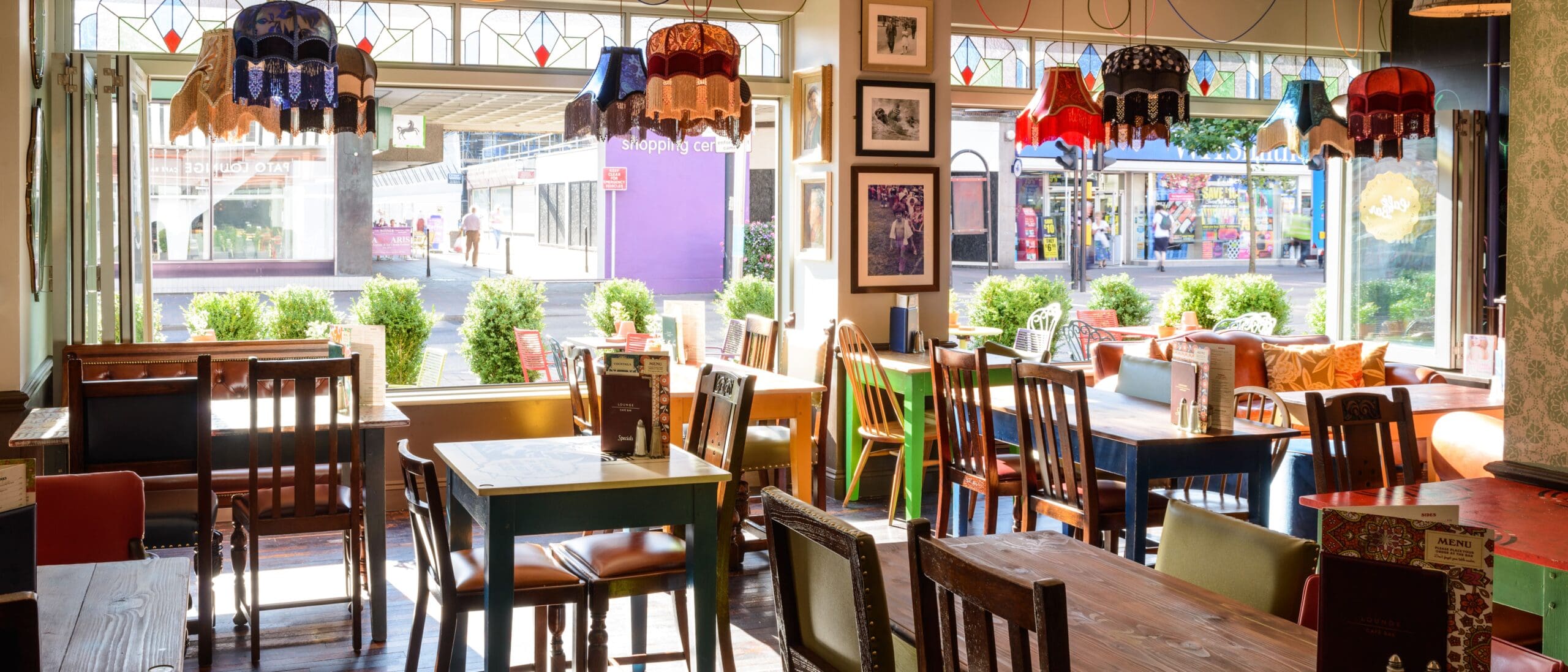 A cozy café with colorful vintage chairs and tables, eclectic lampshades hanging from the ceiling, and large windows letting in sunlight and views of lively Pato street outside.