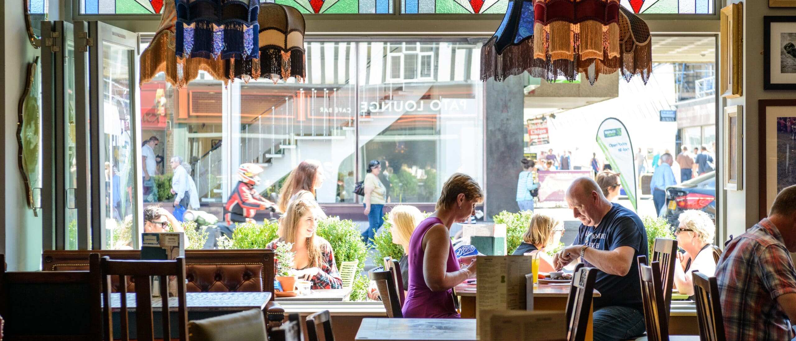 People sit at tables inside cozy Pato café with large windows, sunlight streaming in, and vibrant street life visible outside. Stained glass decorates the top of the window, adding color to the bright scene.