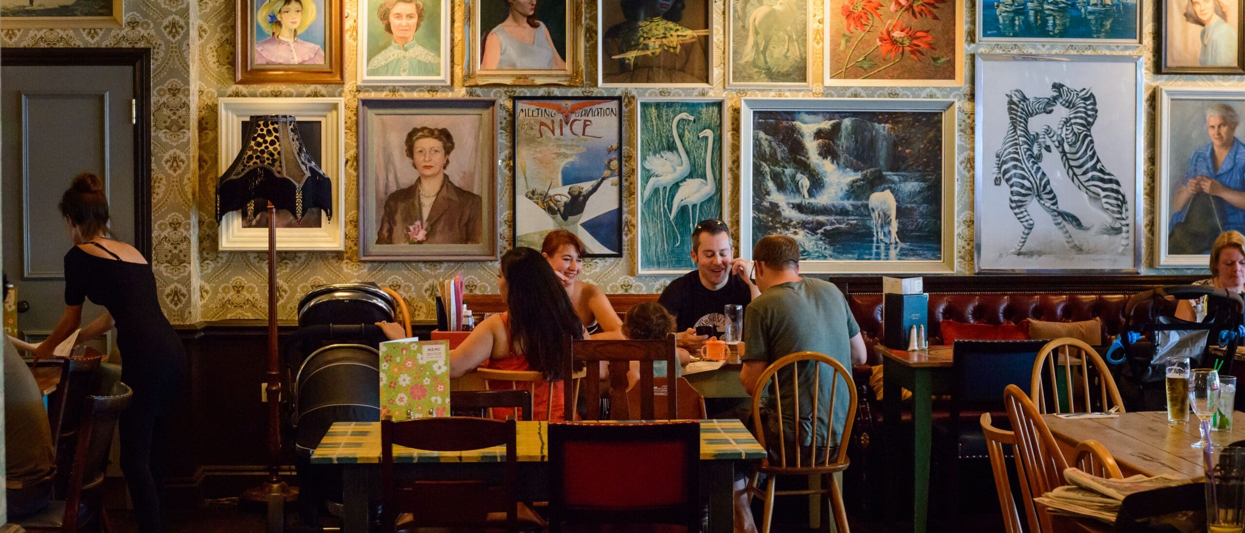 A group of people sit and chat at a table in a cozy café decorated with eclectic framed art, portraits, and a playful painting of Pato, while a server walks by on the left. Other tables and chairs fill the warmly lit space.
