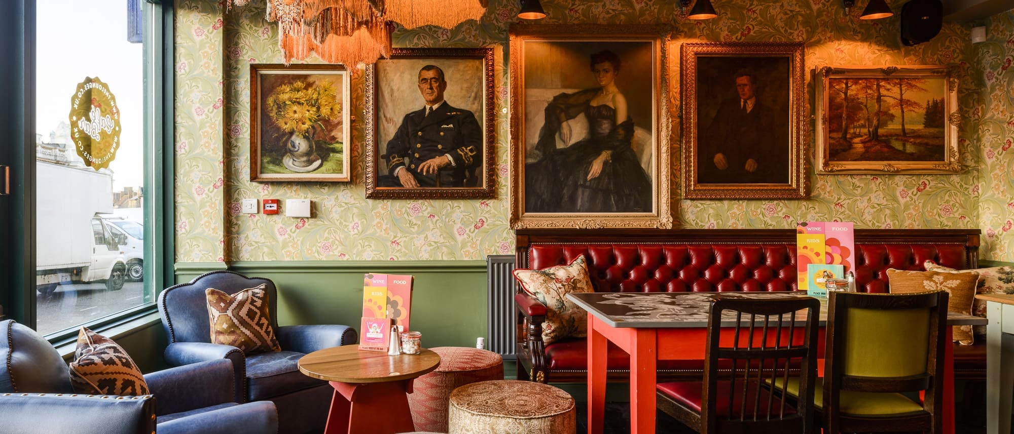 A cozy, vintage-style cafe with velvet seating, floral wallpaper, eclectic cushions, framed paintings reminiscent of the Prado on the wall, and colorful menus on tables near a large window.