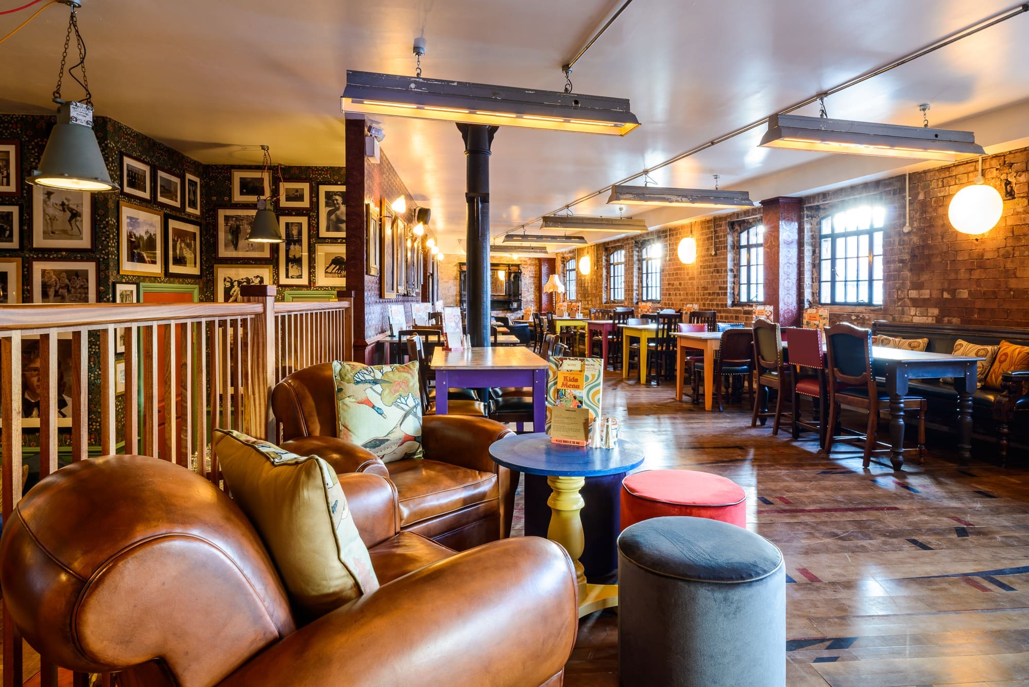 A cozy Puerto pub interior with leather armchairs, wooden floors, framed pictures on brick walls, and tables and chairs arranged along large windows letting in natural light.