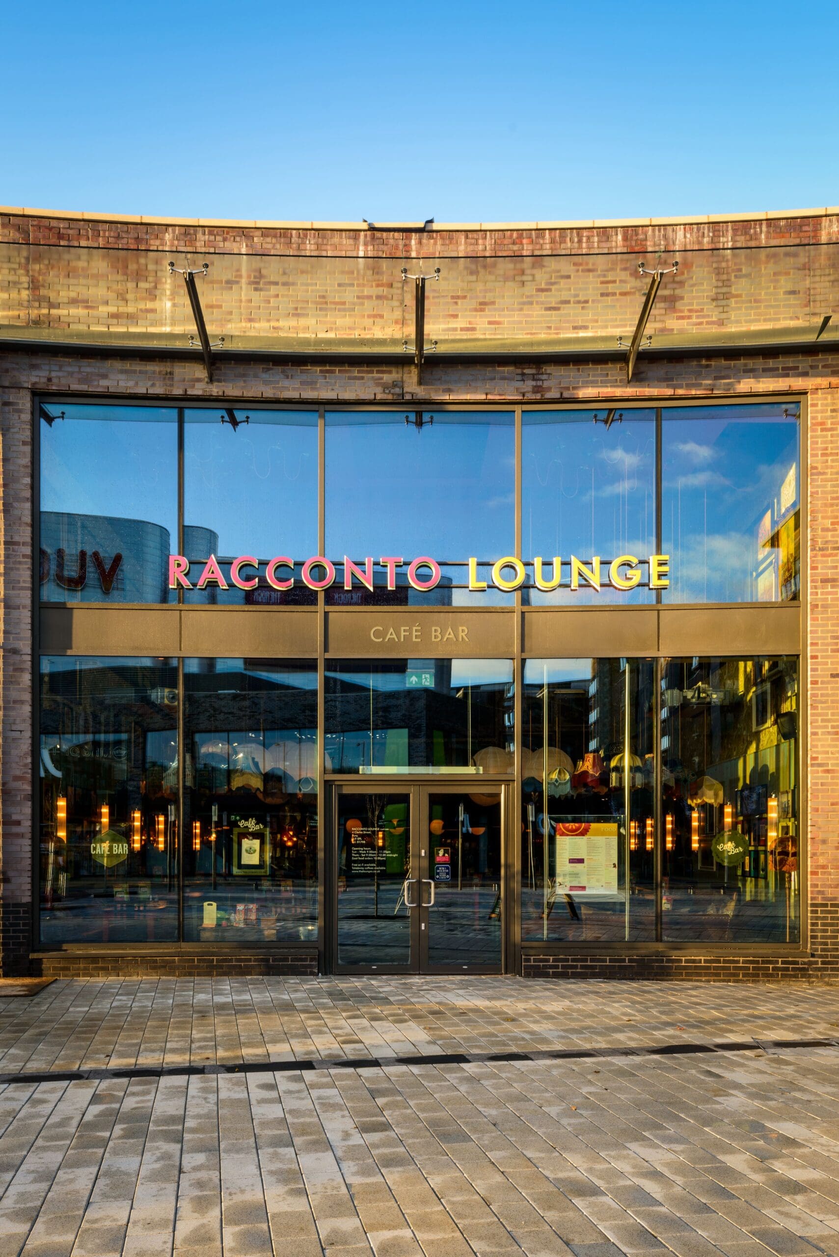 Front view of Racconto Lounge café bar with large glass windows, double doors, and colorful Racconto signage, reflecting the blue sky and surrounding buildings.