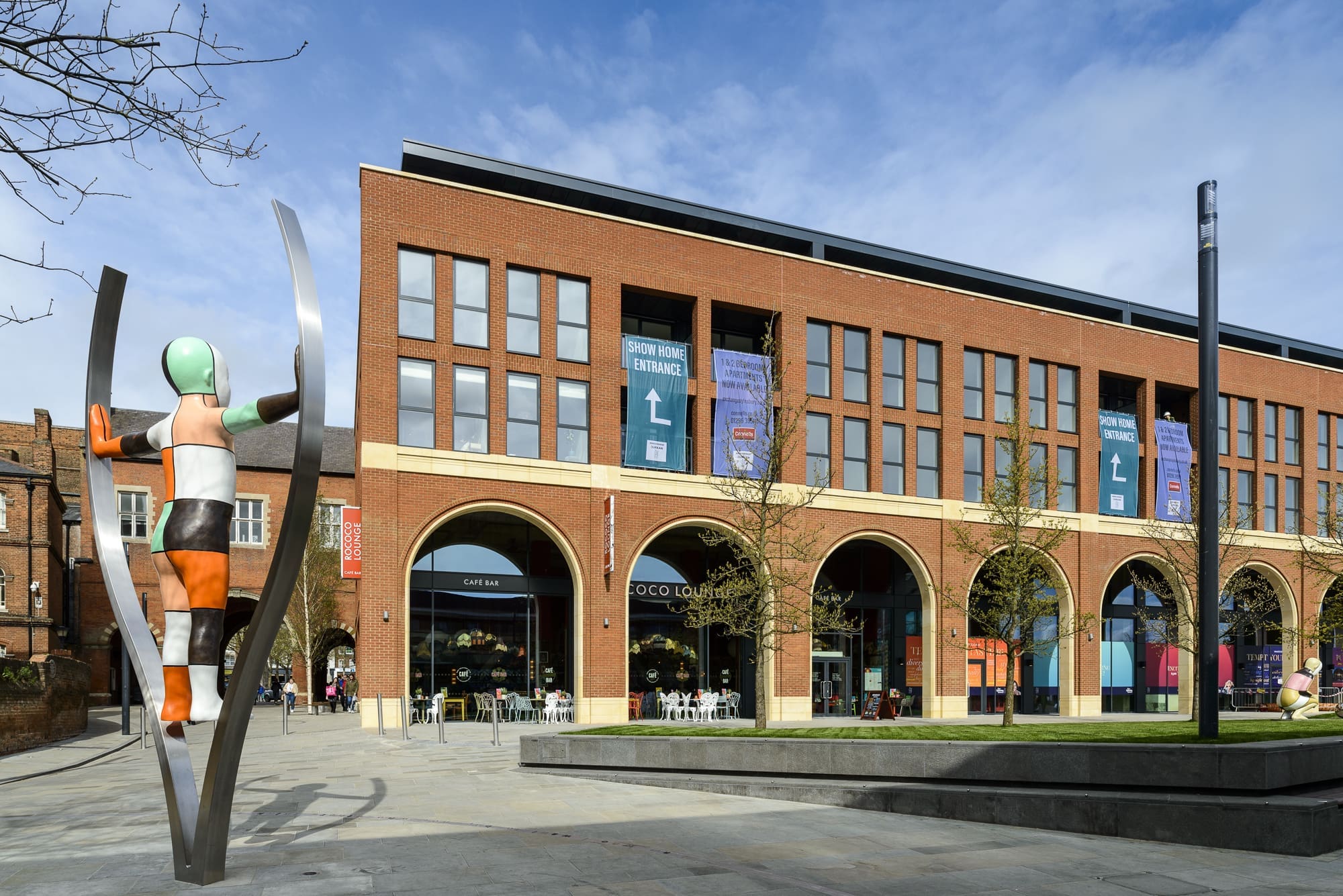A colorful modern sculpture stands in front of a red-brick building with large arched windows, Rococo-style banners, and a café, beneath a blue sky with scattered clouds.