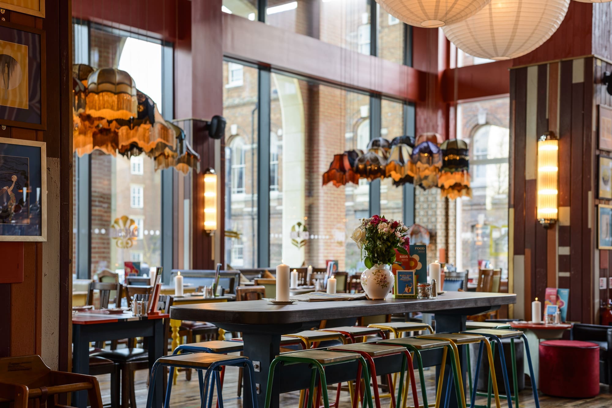 A cozy, colorful café interior features Rococo-inspired eclectic lampshades and mismatched chairs around a long table with flowers in a vase. Soft lighting and urban views create a warm, inviting atmosphere.